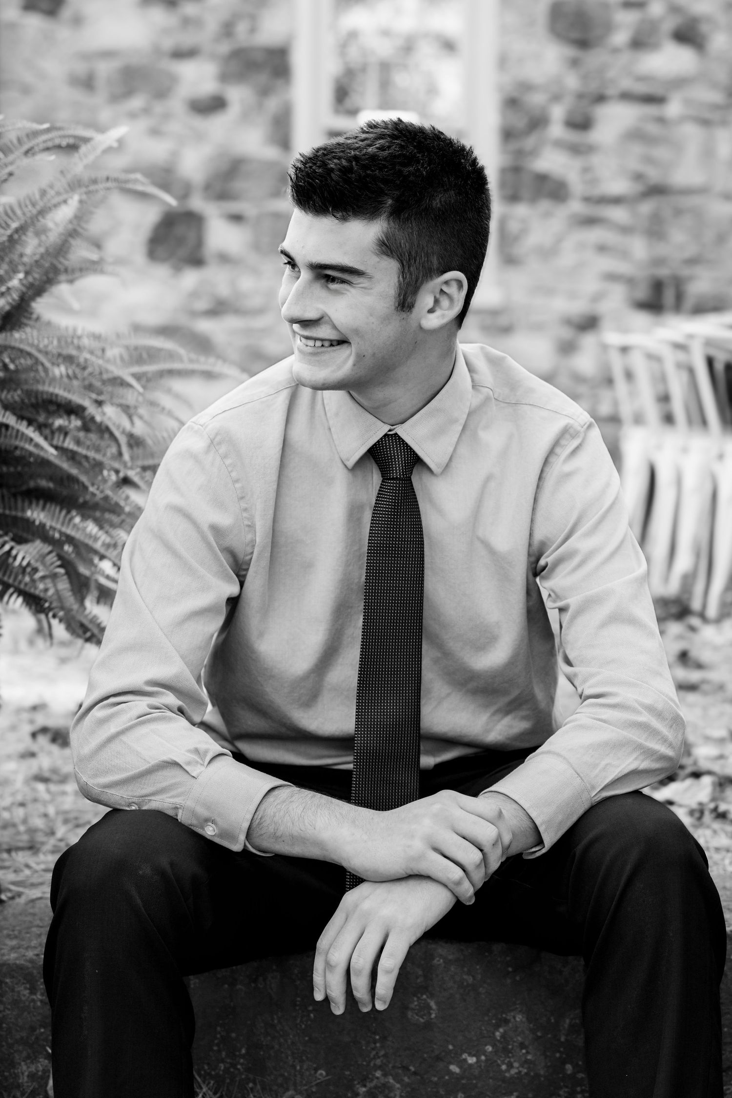 Black and white portrait series showing a high school senior young man in business attire sitting and smiling in an outdoor setting.