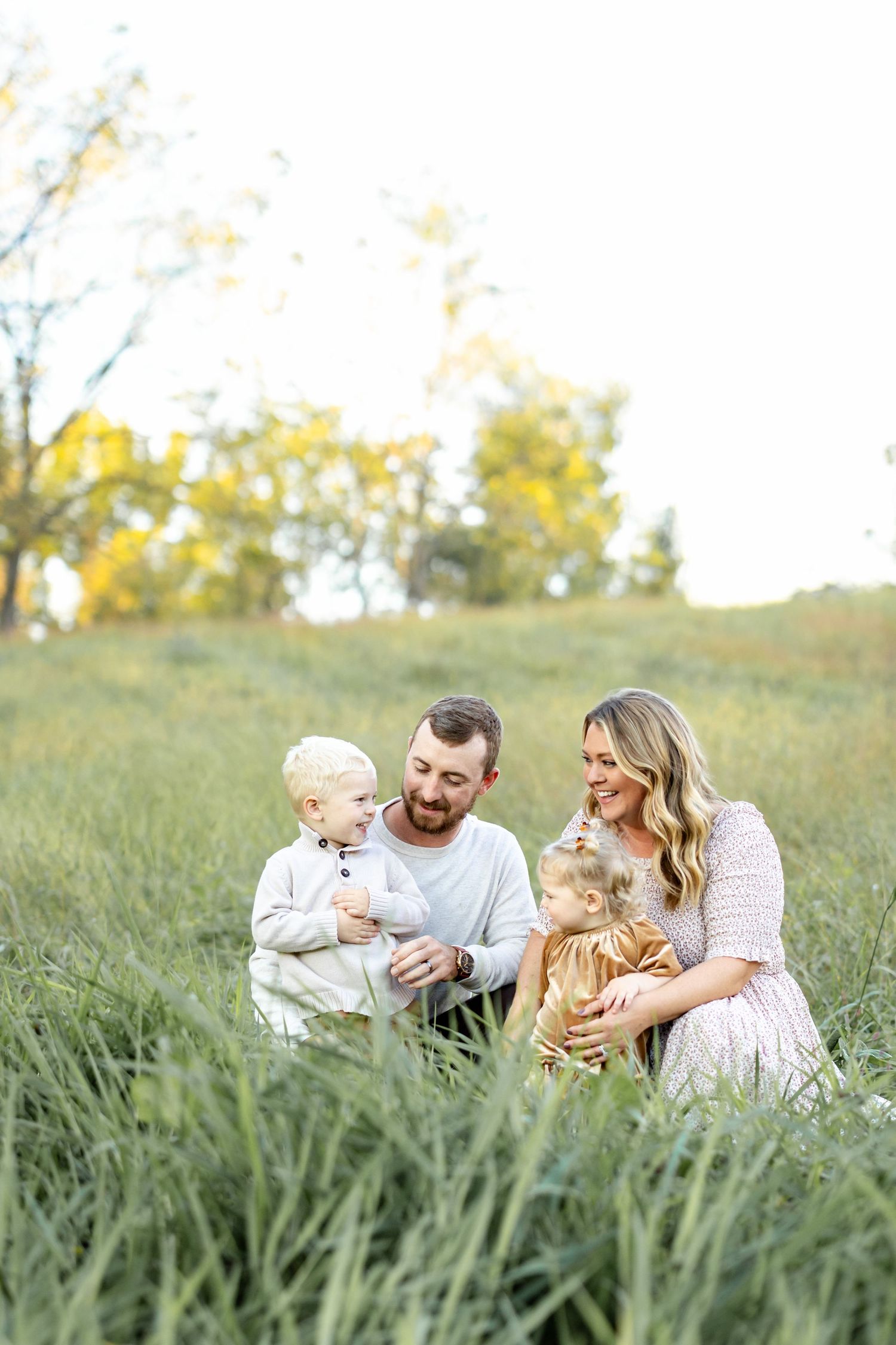 A family relaxes together in a grassy meadow on a sunny day with golden trees in the background.