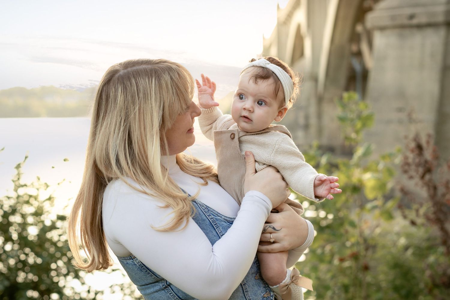 A blonde mother tenderly holds her baby girl outdoors near a bridge, bathed in warm golden sunlight.
