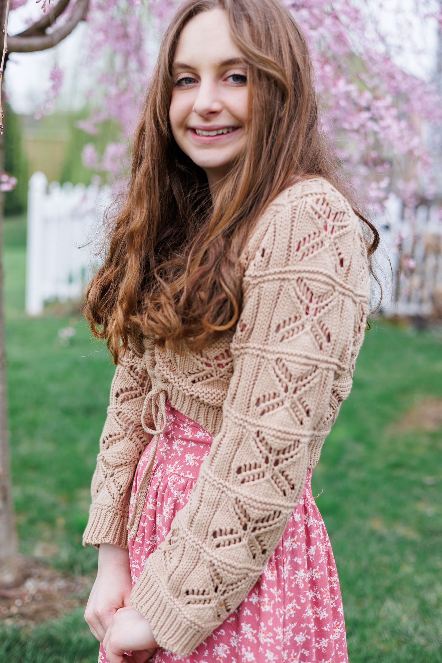 Young person graduating high school in pink floral dress and beige knit cardigan poses outdoors near blooming cherry blossom trees.