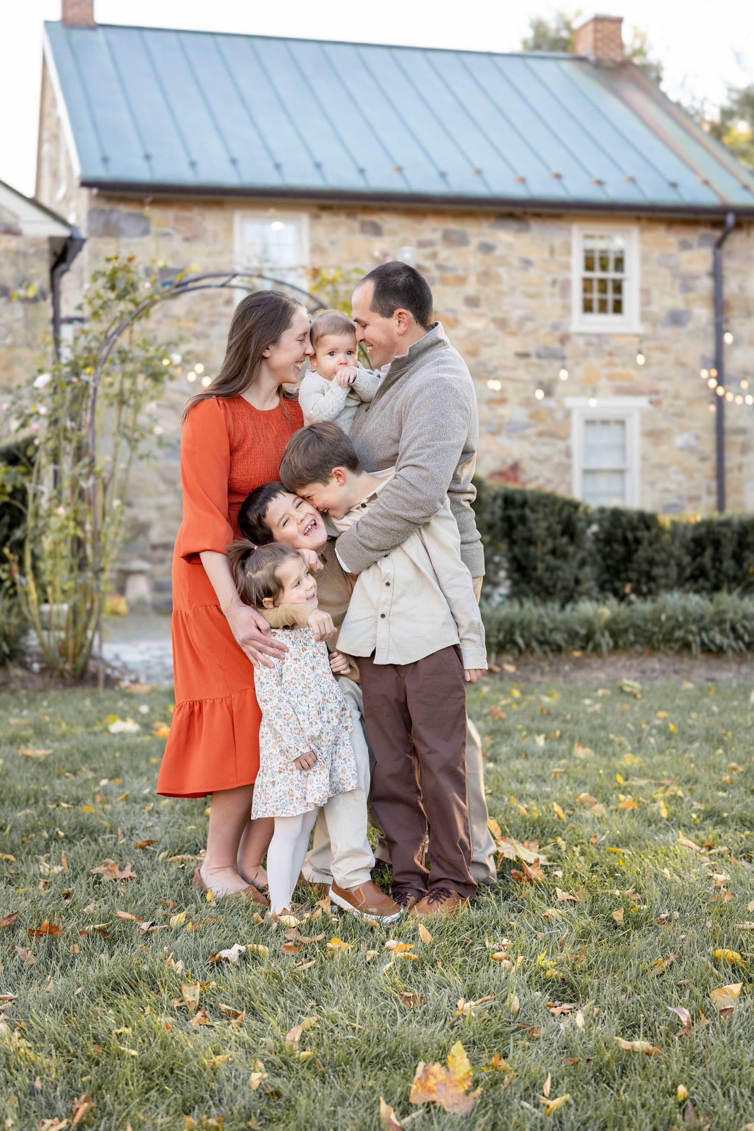 A happy family of five poses outdoors in fall, parents holding children, with a stone house backdrop.