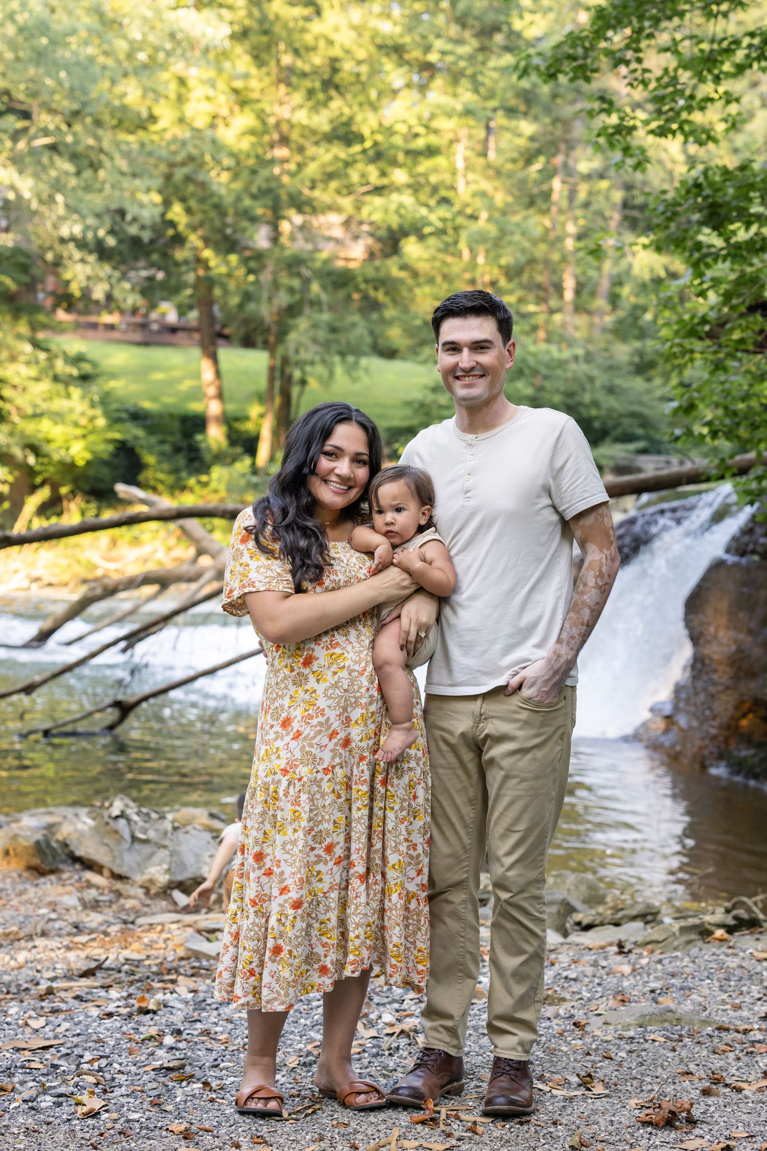 A family with their baby poses together at a scenic waterfall location in Lancaster County, PA, surrounded by lush greenery on a sunny day.