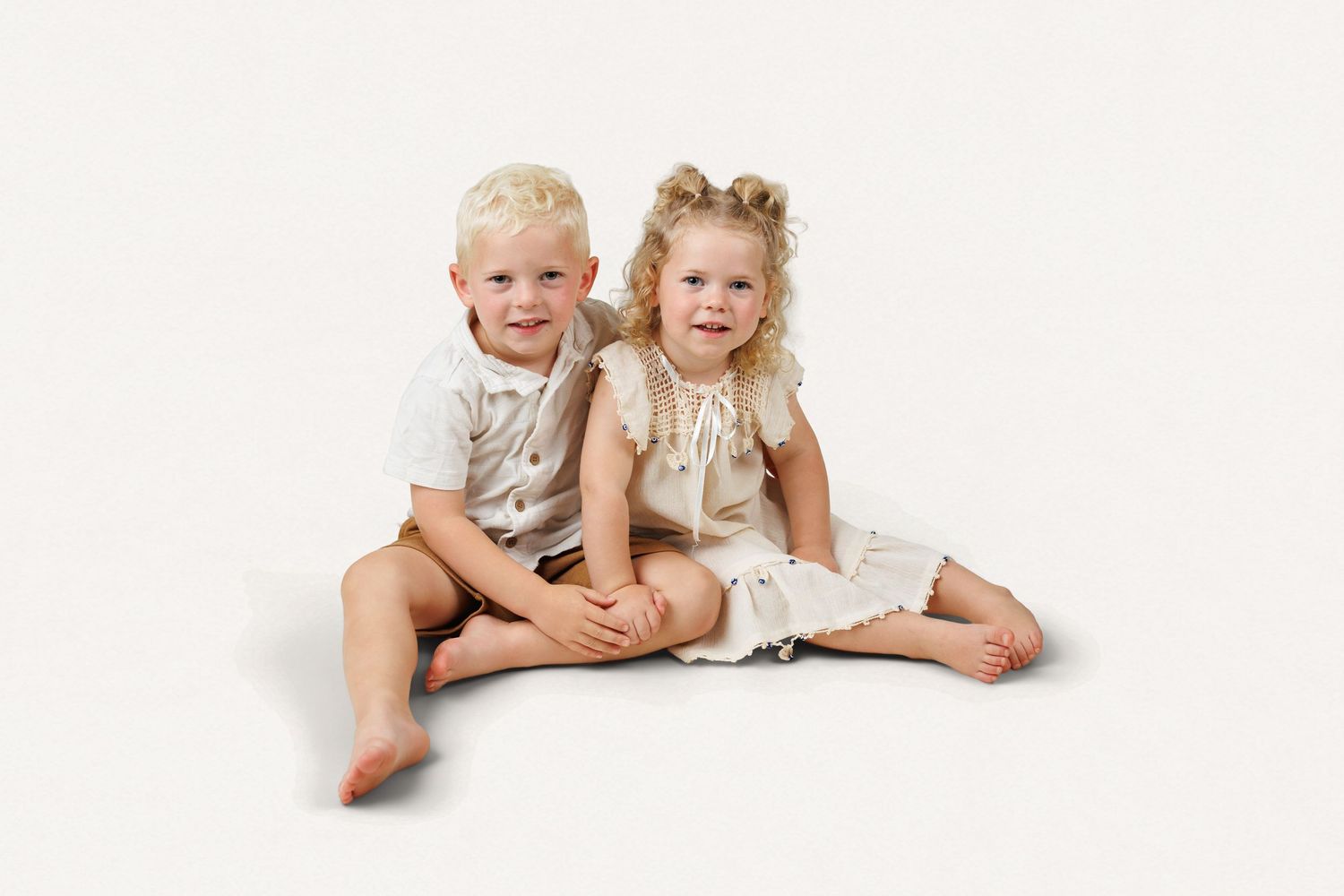 Two blonde toddlers in cream outfits sit together smiling on a white background.