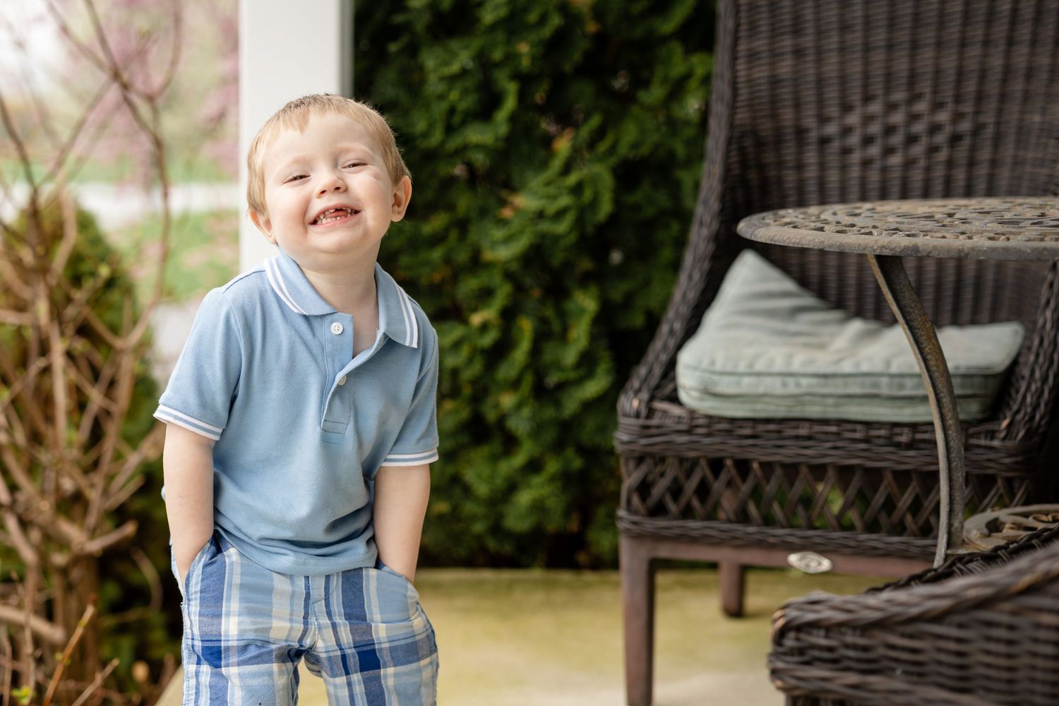 Happy toddler boy in blue polo shirt and plaid shorts laughing outdoors on a patio with wicker furniture.