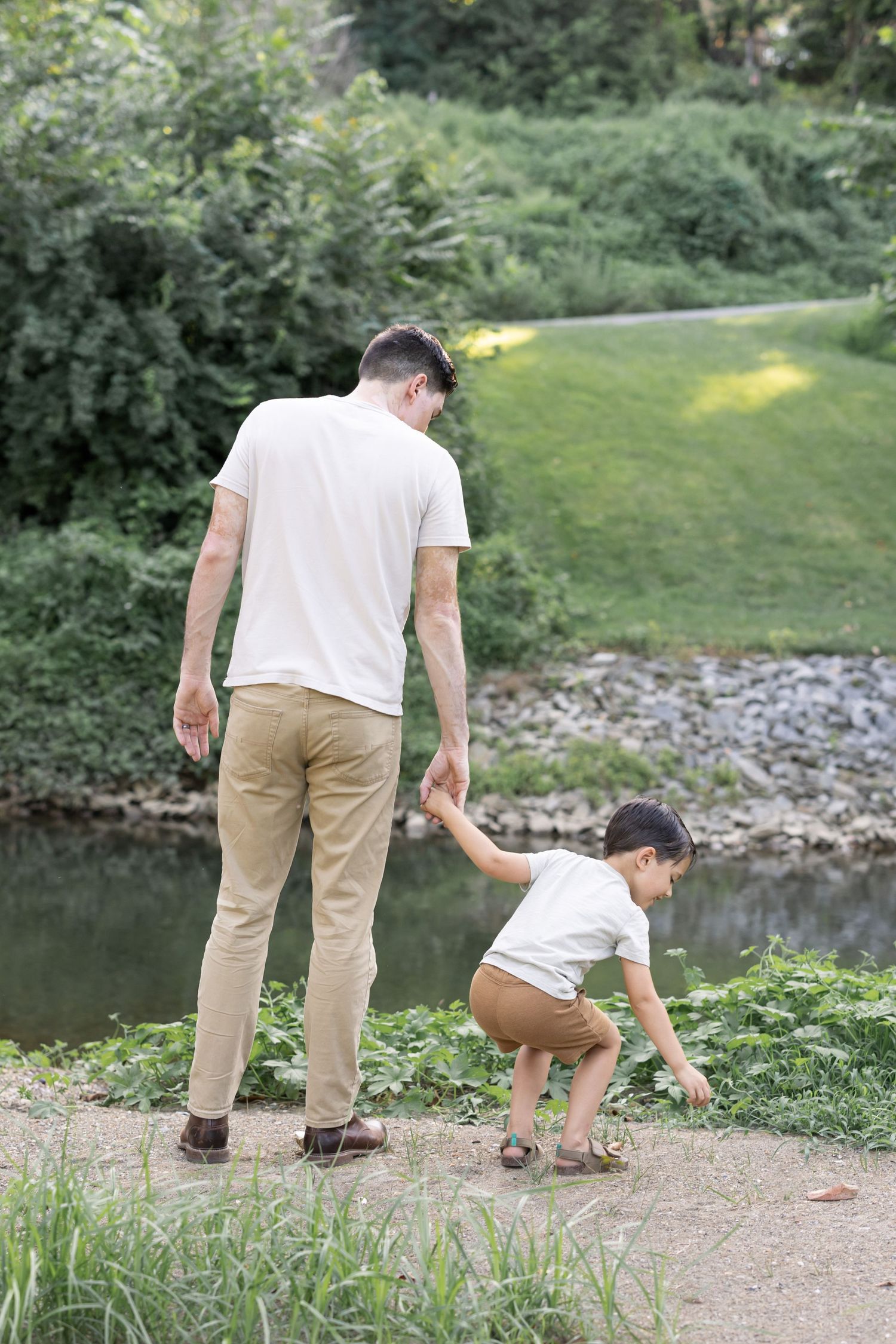 Couple crouches by a riverbank on a summer afternoon searching for stones in front of lush green foliage during family photos.