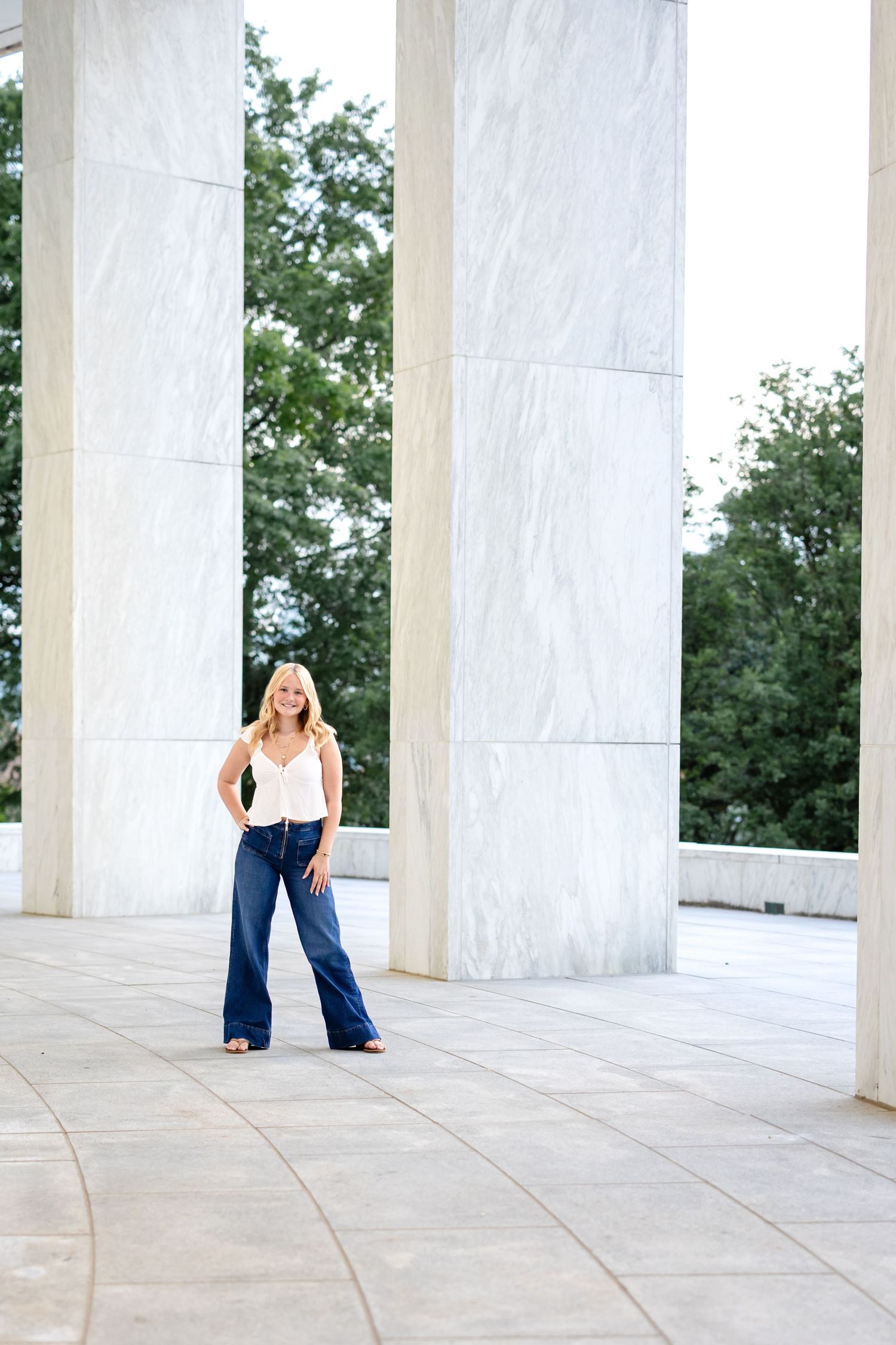 A high school senior girl in a white top and blue pants poses between marble columns in an outdoor architectural setting.