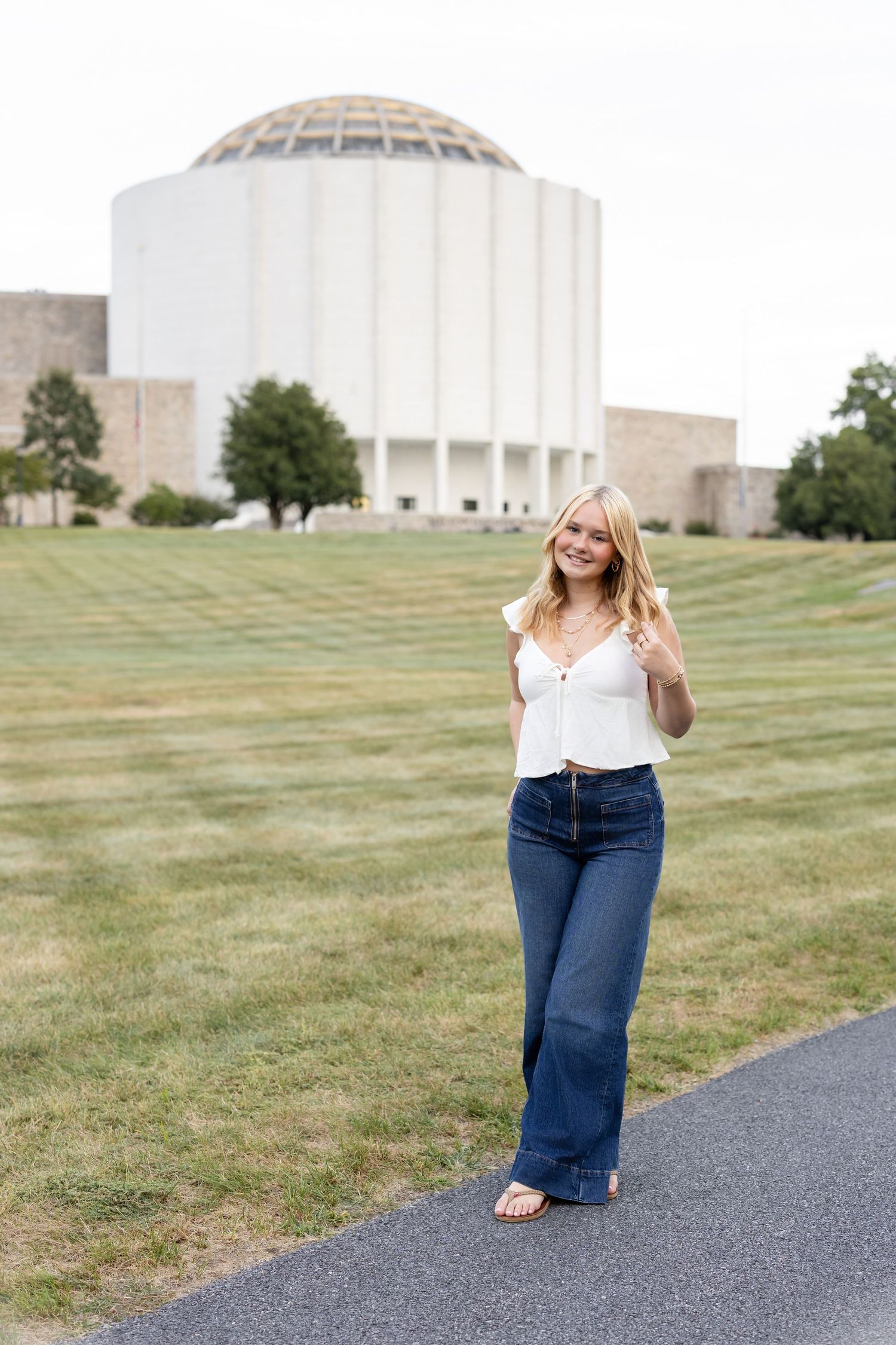 A high school senior girl in white top and flared jeans stands on a path in front of a large domed building on a grassy campus.