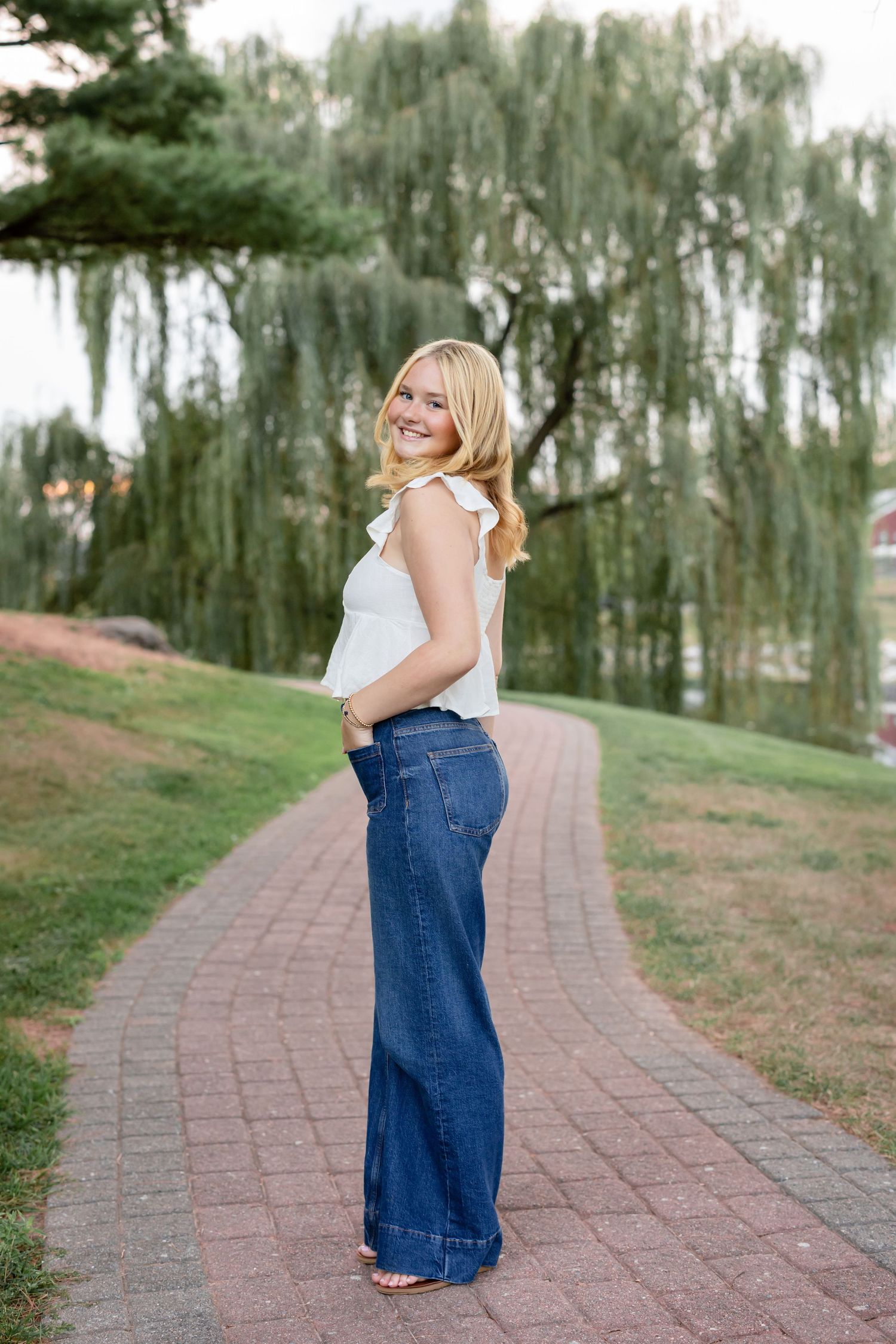 A figure in a white top and jeans walks along a curved brick path bordered by willow trees.