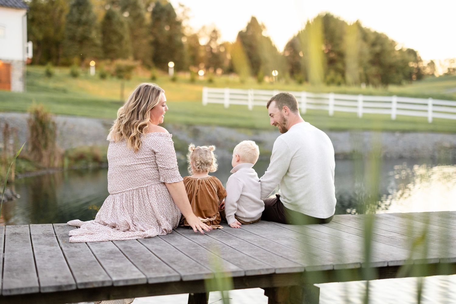 Family of four sitting on a wooden dock by a pond at golden hour, enjoying a serene countryside view.