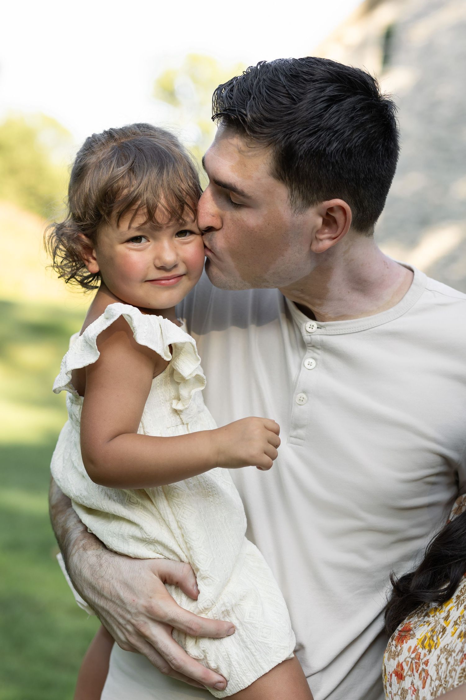 A tender moment as a parent gives a loving kiss on the cheek to a young child wearing a white dress outdoors in Lancaster, PA.