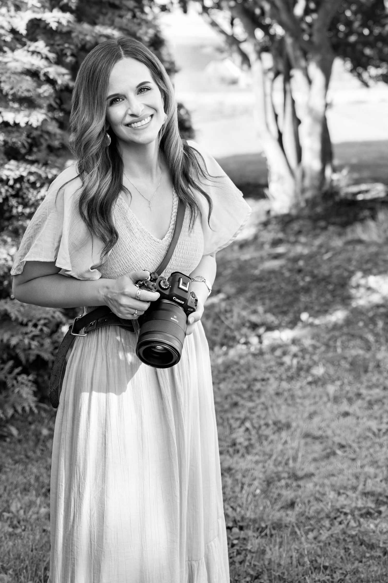 Black and white photo of a smiling woman holding a DSLR camera outdoors in a garden setting.