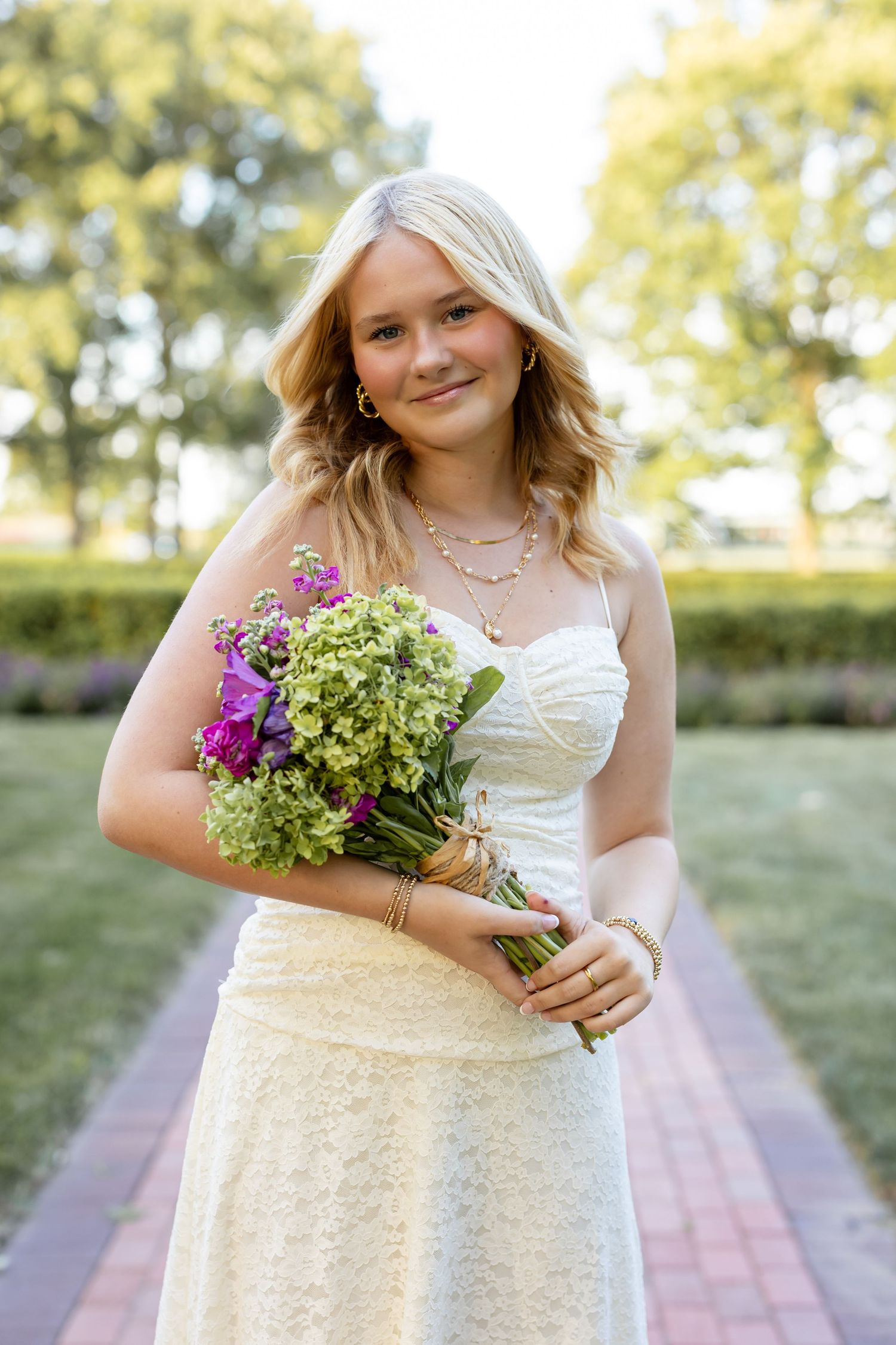 A high school senior girl in a white wedding dress holds a beautiful bouquet of green hydrangeas and purple flowers outdoors.