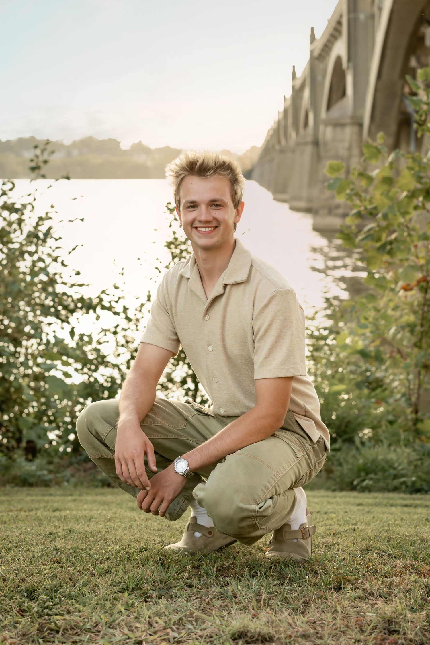 A casual outdoor portrait session featuring a young man in beige clothing crouching on grass near water at sunset.
