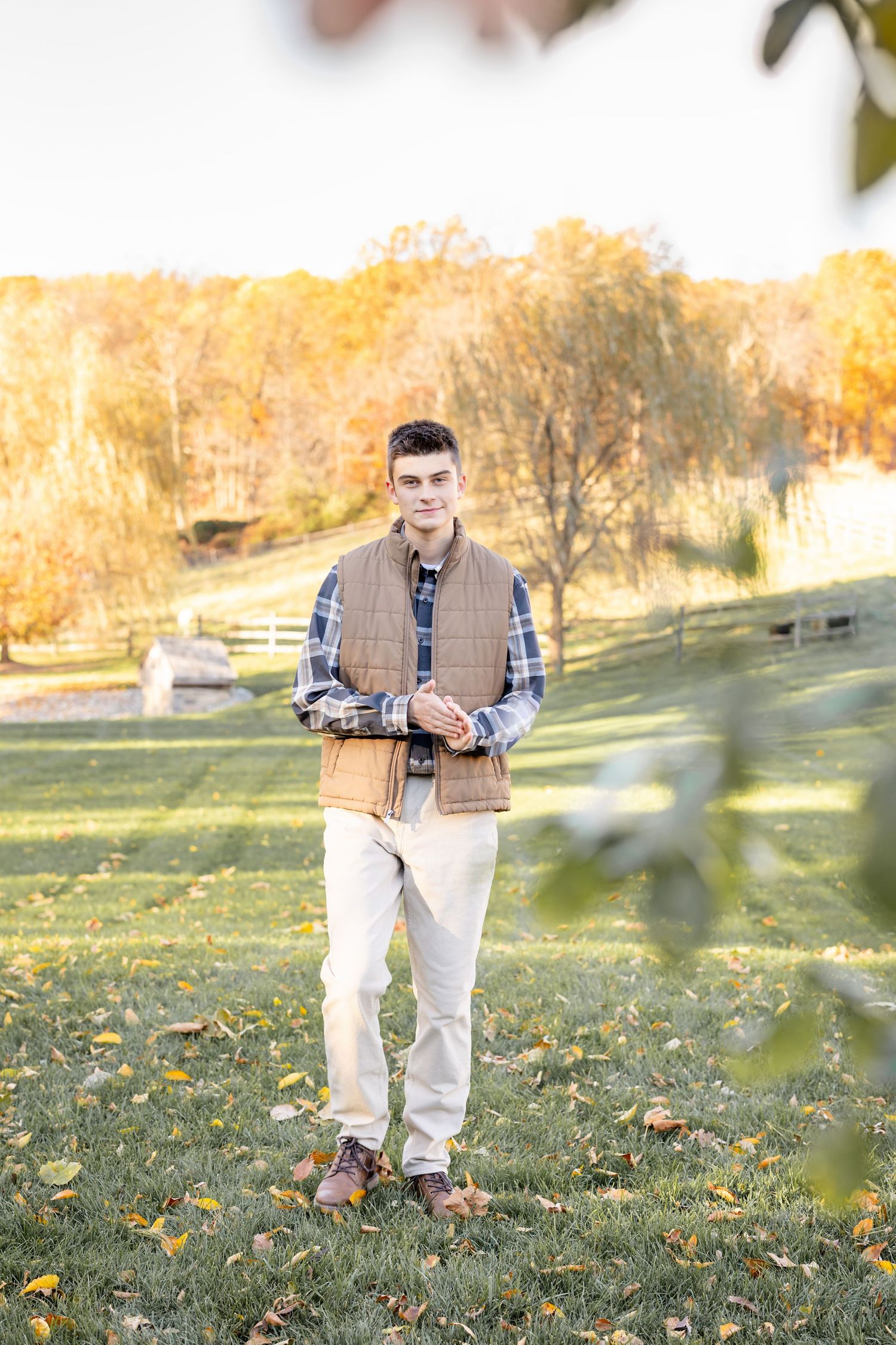Person wearing plaid shirt and brown vest poses in grassy autumn field at golden hour.