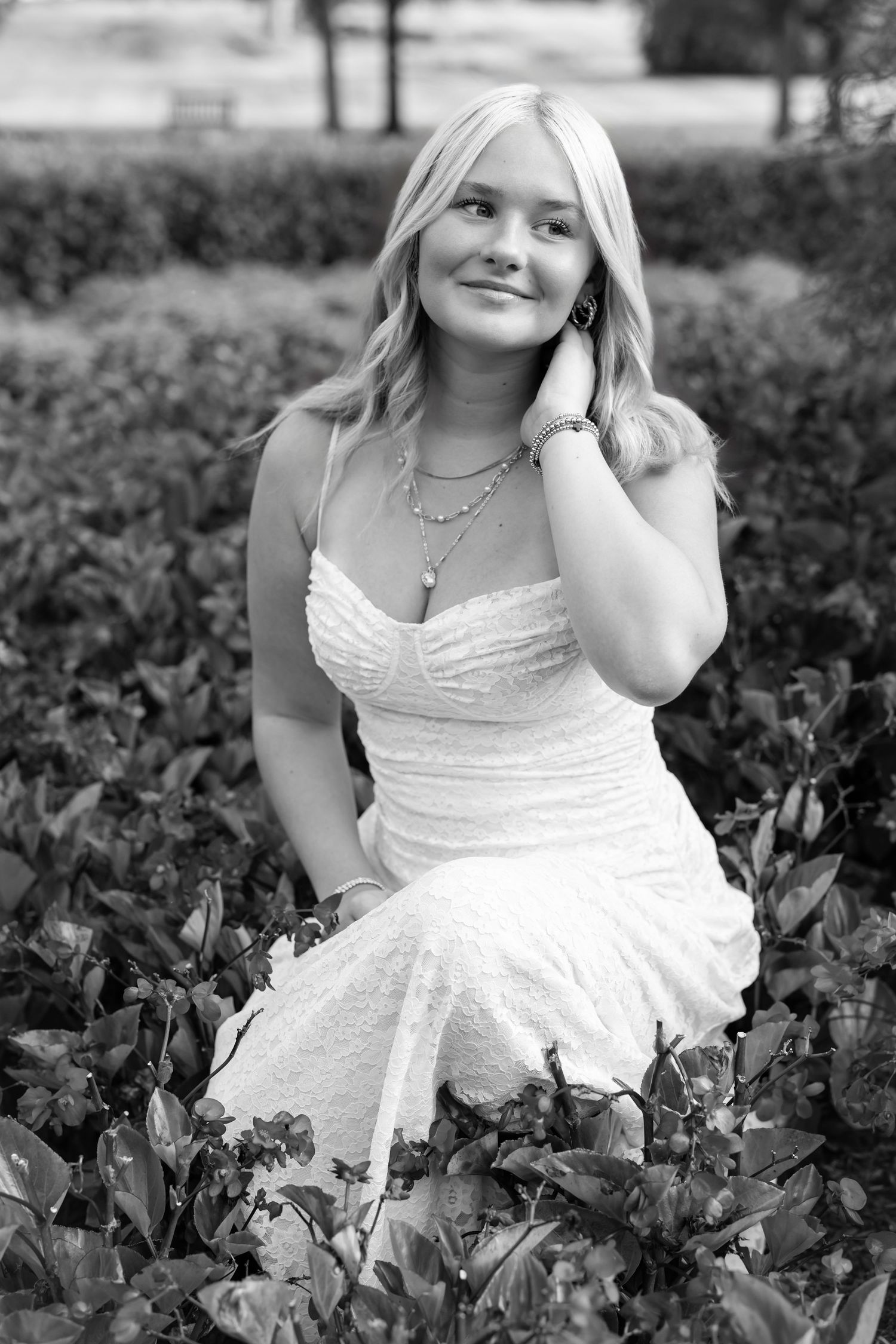 Black and white portrait series showing a high school graduate in a white dress sitting among garden foliage.