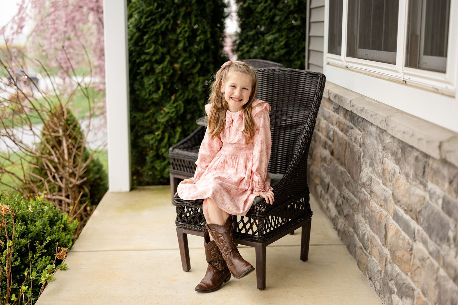 Young girl in pink dress and brown cowboy boots sitting on a wicker chair on a stone porch.