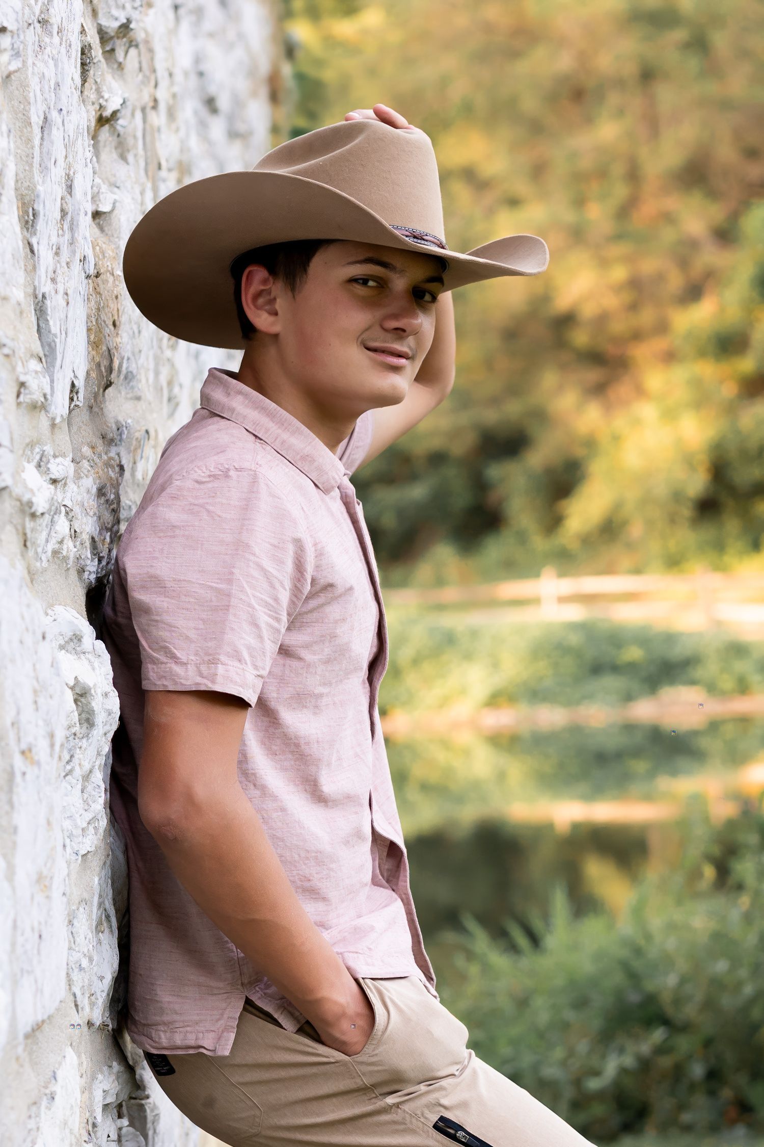 A high school senior boy leans against a rustic stone wall wearing a faded red button down and a brown cowboy hat for his senior photos.