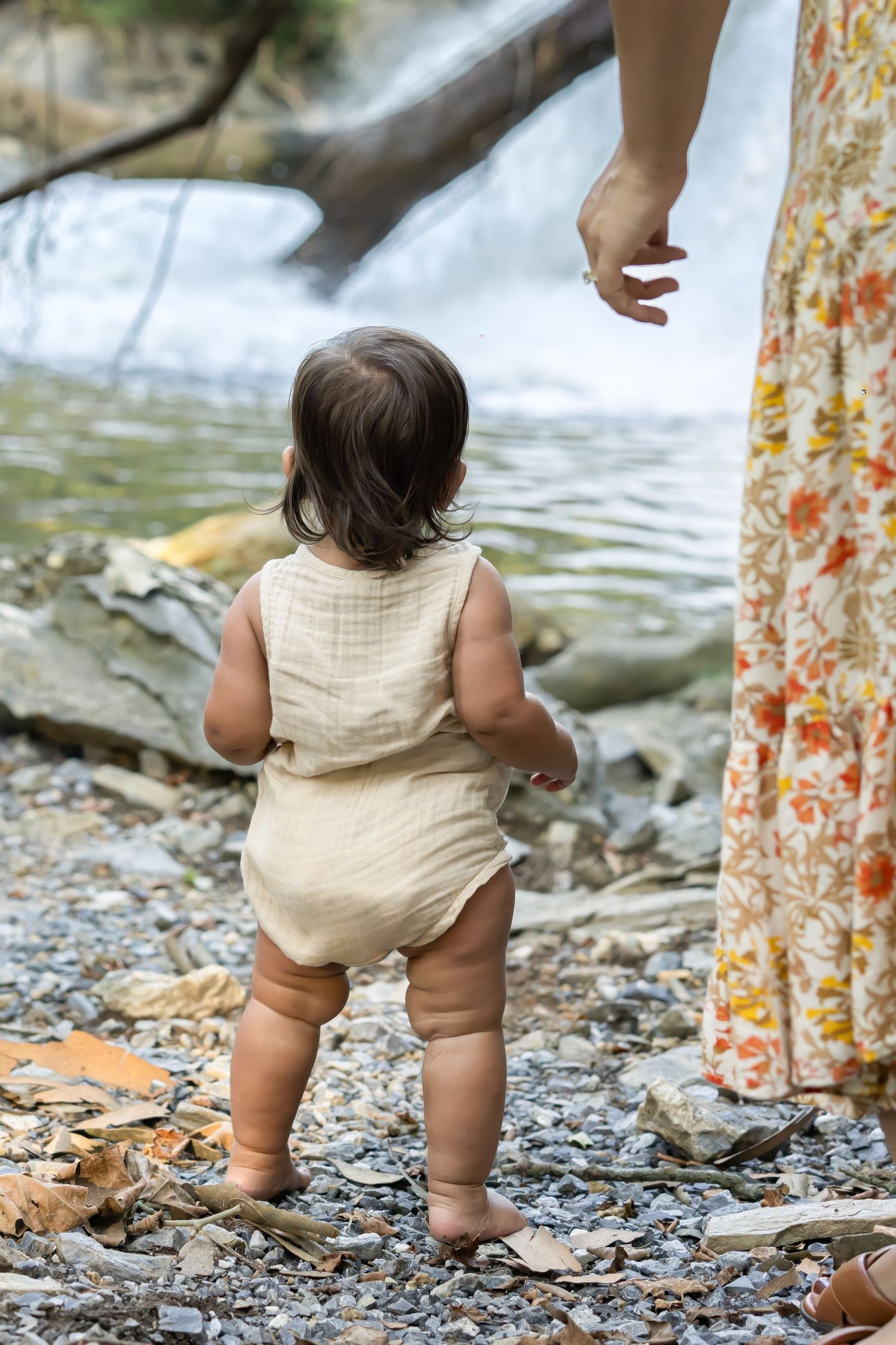 Small child in beige romper stands on rocky ground near flowing water in Lancaster, PA while watched by adult in floral dress.
