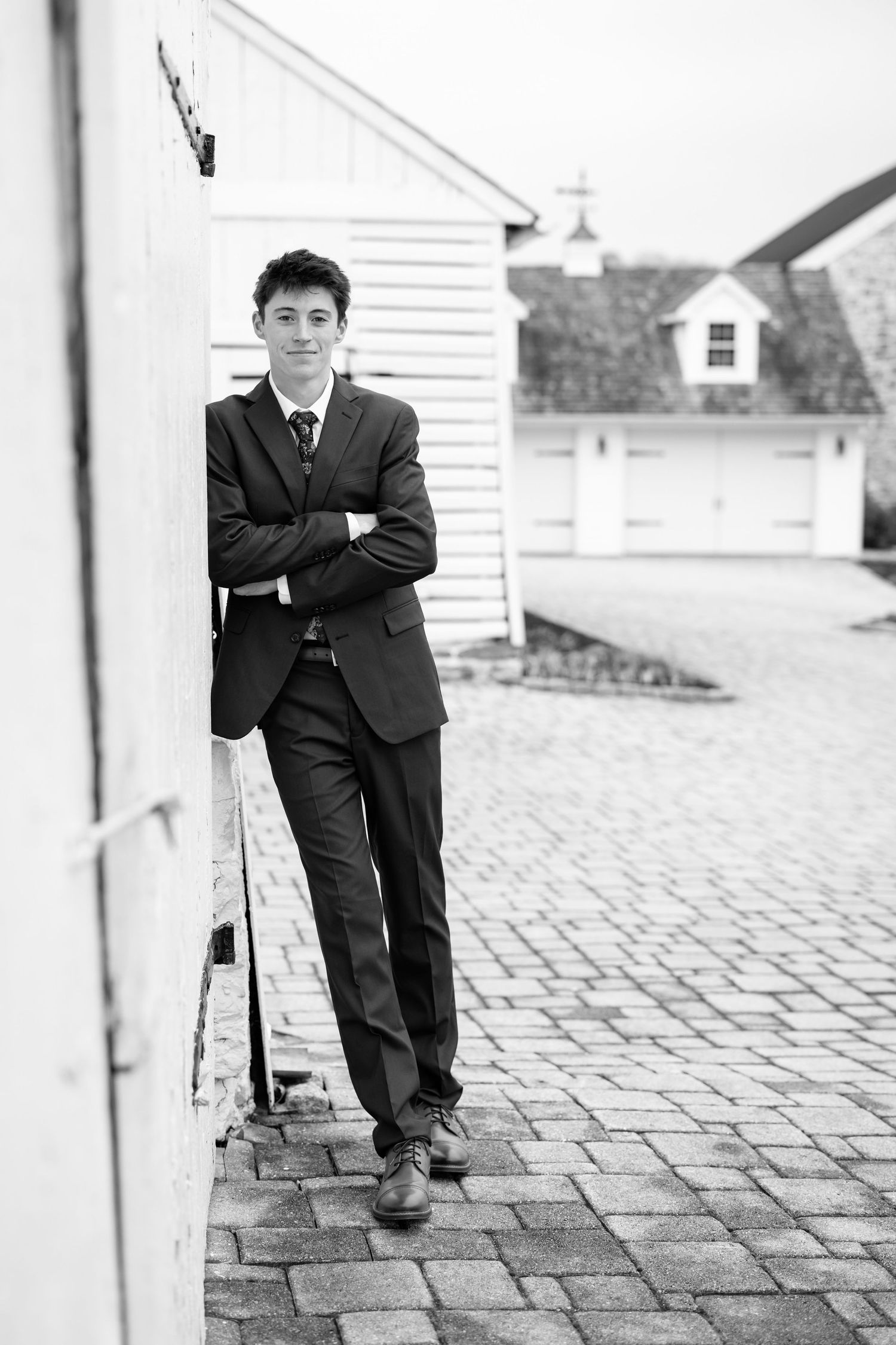 Black and white photo of young man in a suit leaning against a white post on a brick patio | luxury senior portraits in Lancaster, PA.