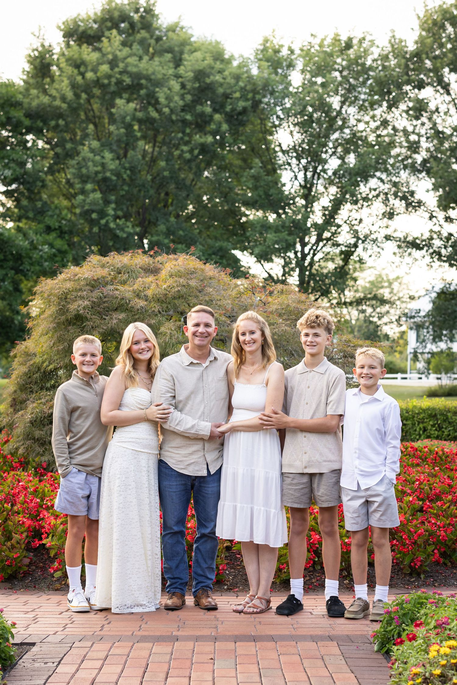 Family group poses together around a graduate in coordinated white and neutral outfits against a colorful garden backdrop with red flowers.