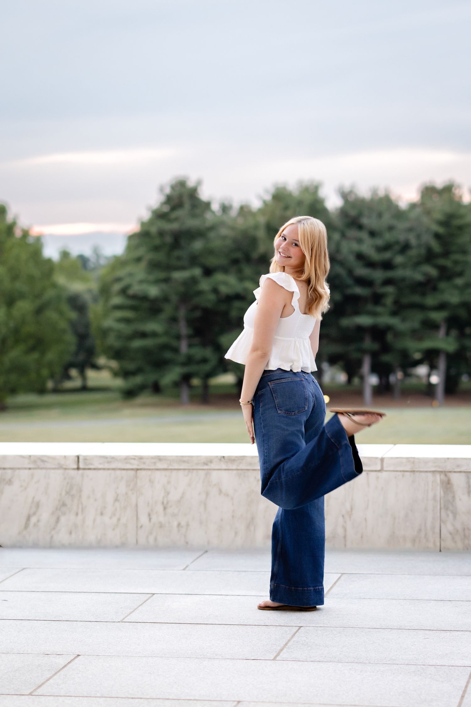 A high school senior girl in white sleeveless top and wide-leg jeans poses on concrete ledge with evergreen trees in background.
