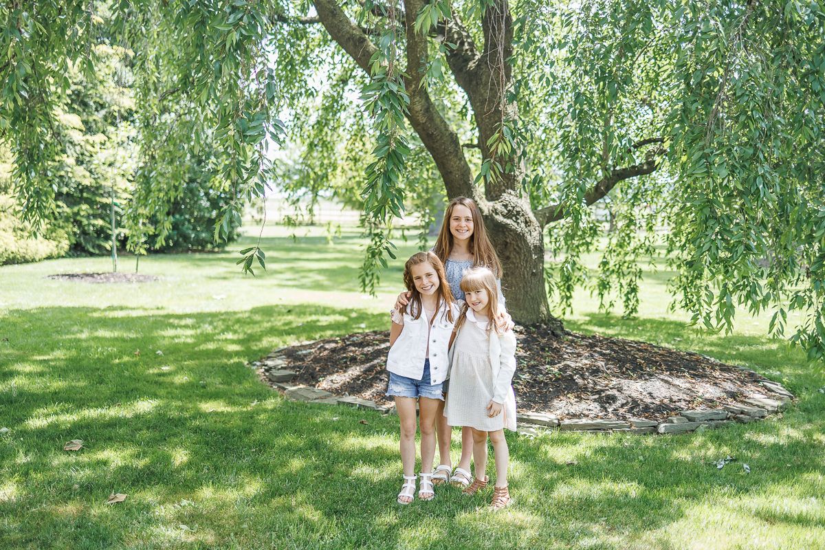 Megan Mariah Photography shows three sisters posing under willow tree at White Chimney Estates in Gap, PA during family portraits.