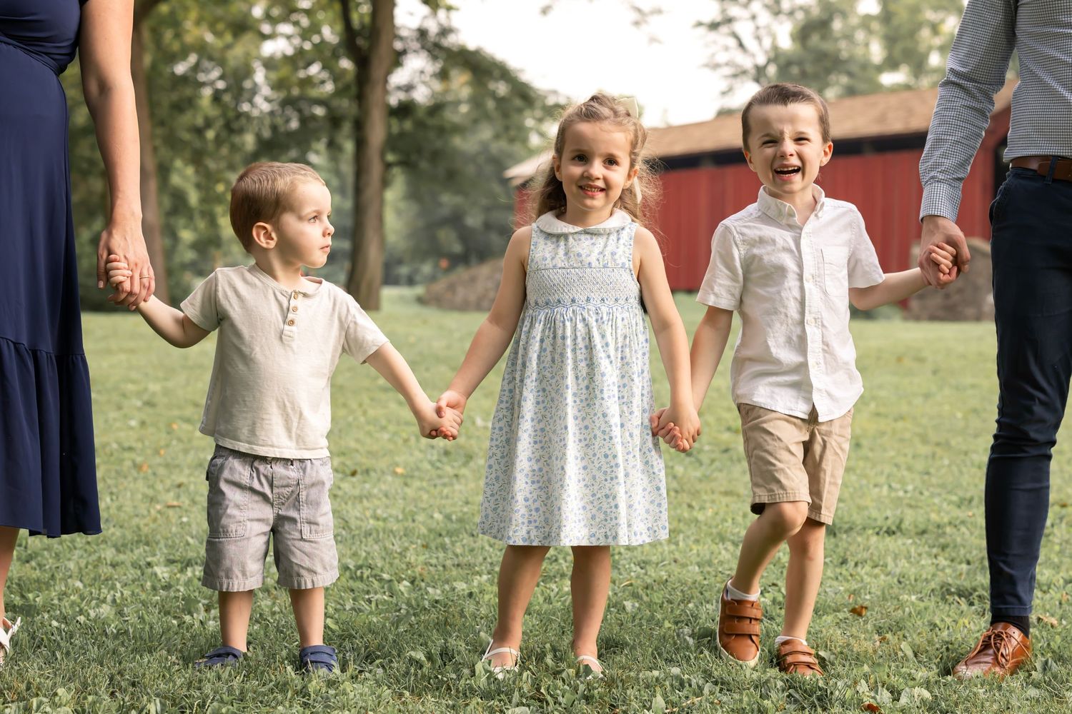 Three young children holding hands and smiling outdoors on green grass with a red barn in the background.