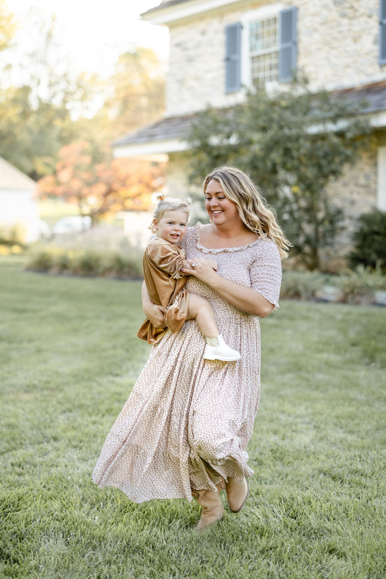 A smiling woman in a floral dress holds a happy toddler while walking on green grass in front of a stone house.