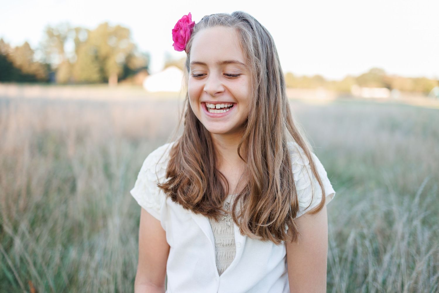 A joyful young girl with long brown hair and a pink flower accessory laughs in a sunlit field wearing a white dress.