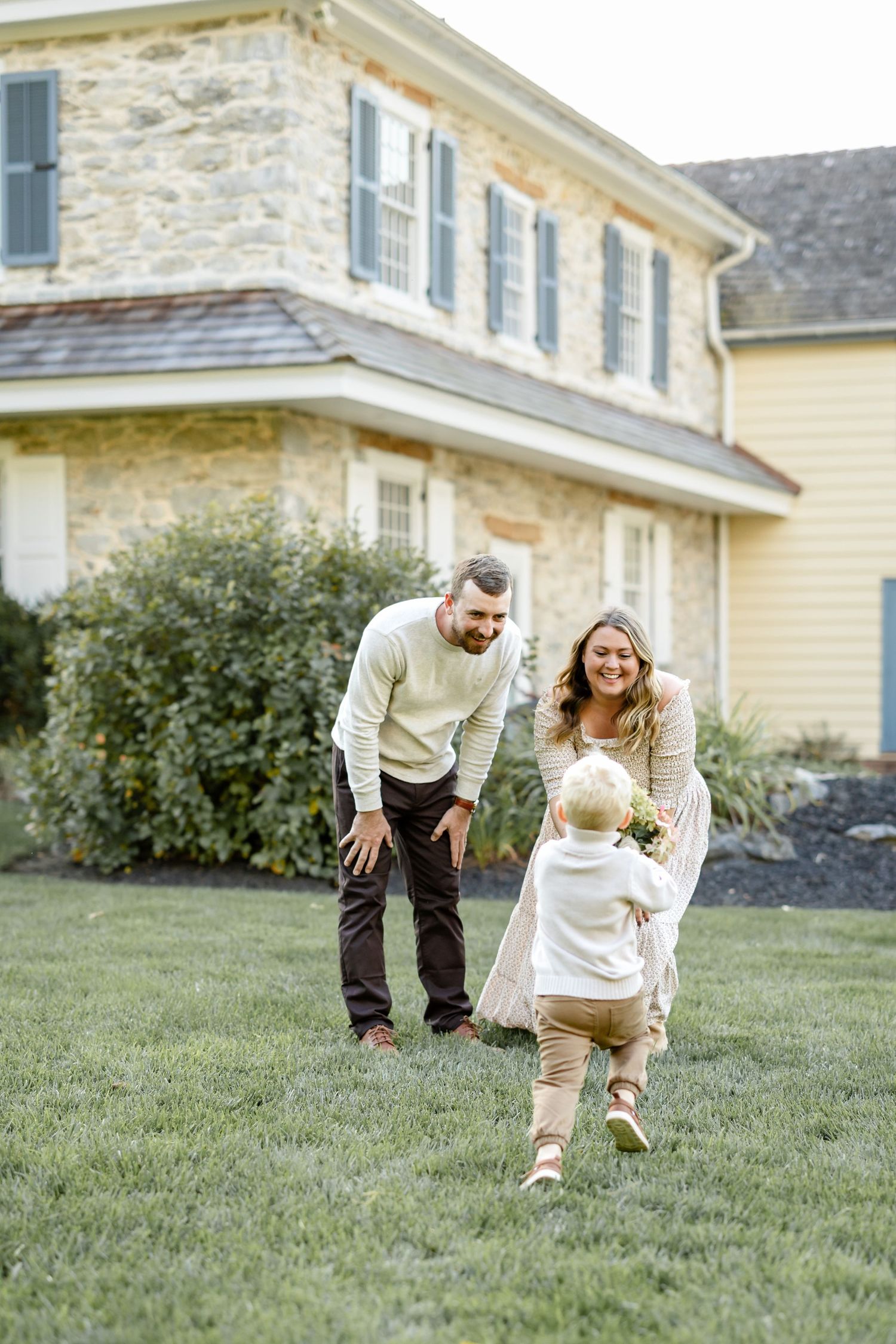 A happy family with a toddler playing on green grass in front of a charming stone and yellow farmhouse.
