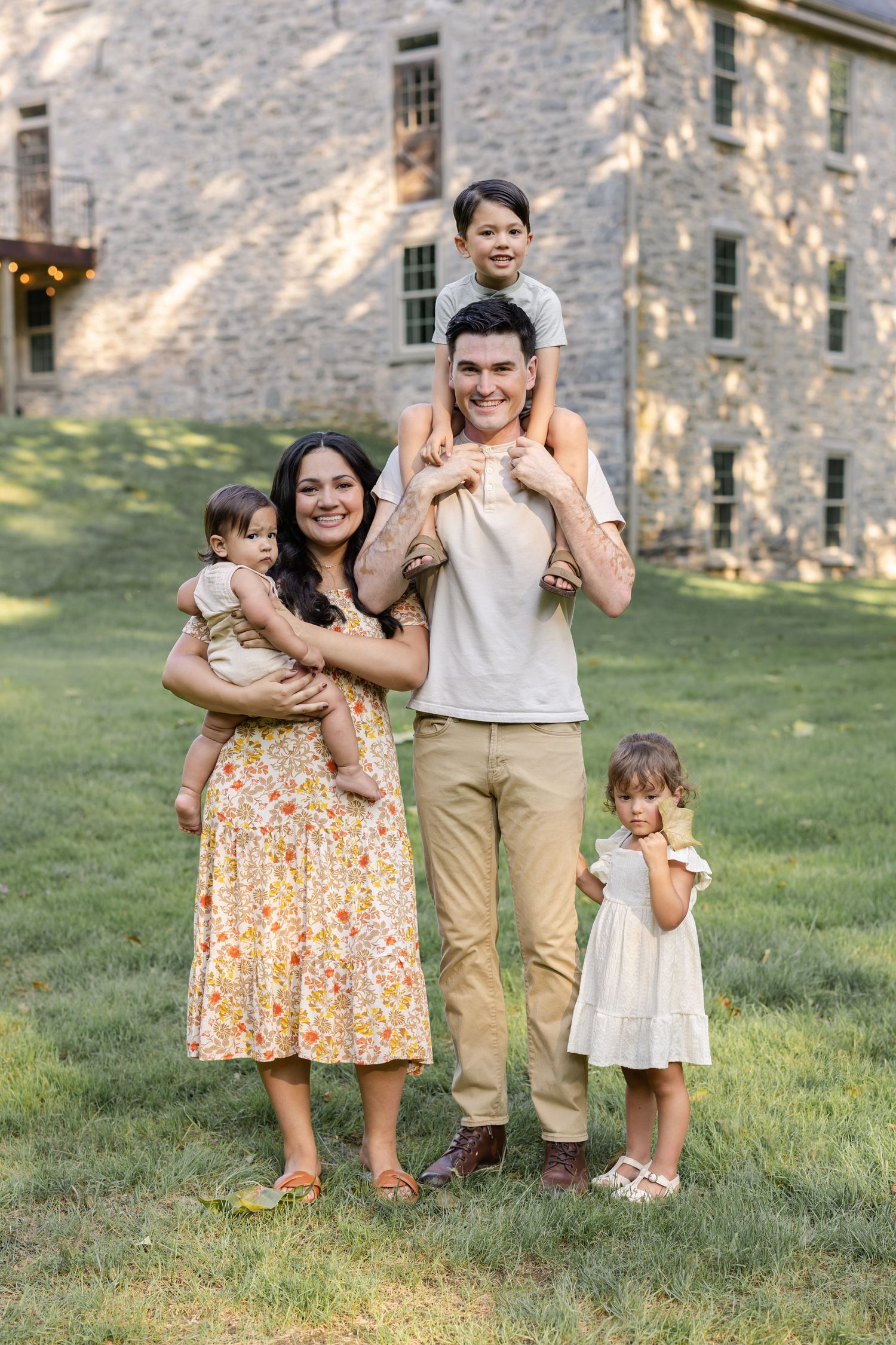 A smiling family of five poses outdoors on green grass in front of a stone building on a sunny day.