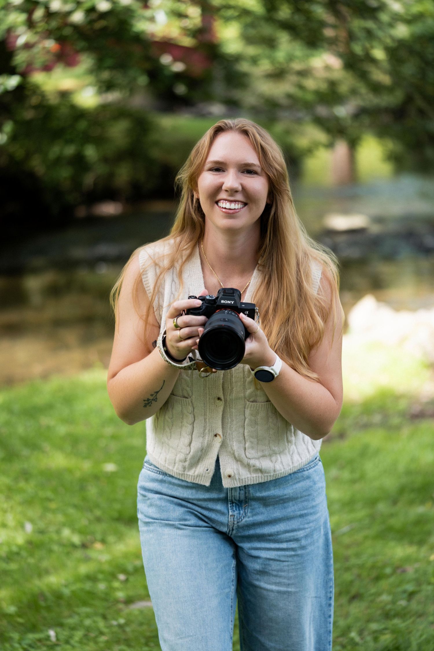 A photographer holding a camera stands outdoors in jeans and a vest against a natural green background.