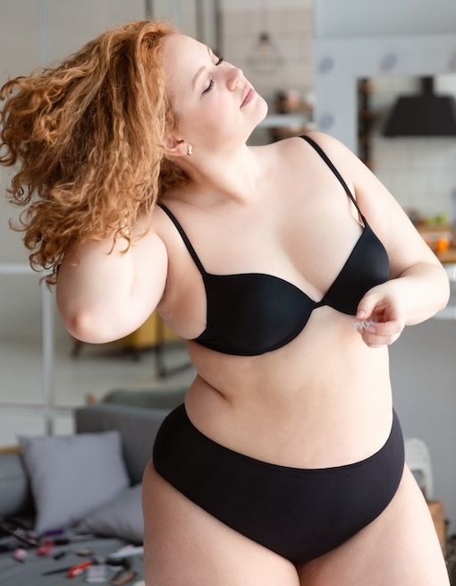 Confident woman with curly red hair in black lingerie flipping her hair while getting ready for a boudoir session in a natural, lived-in spa