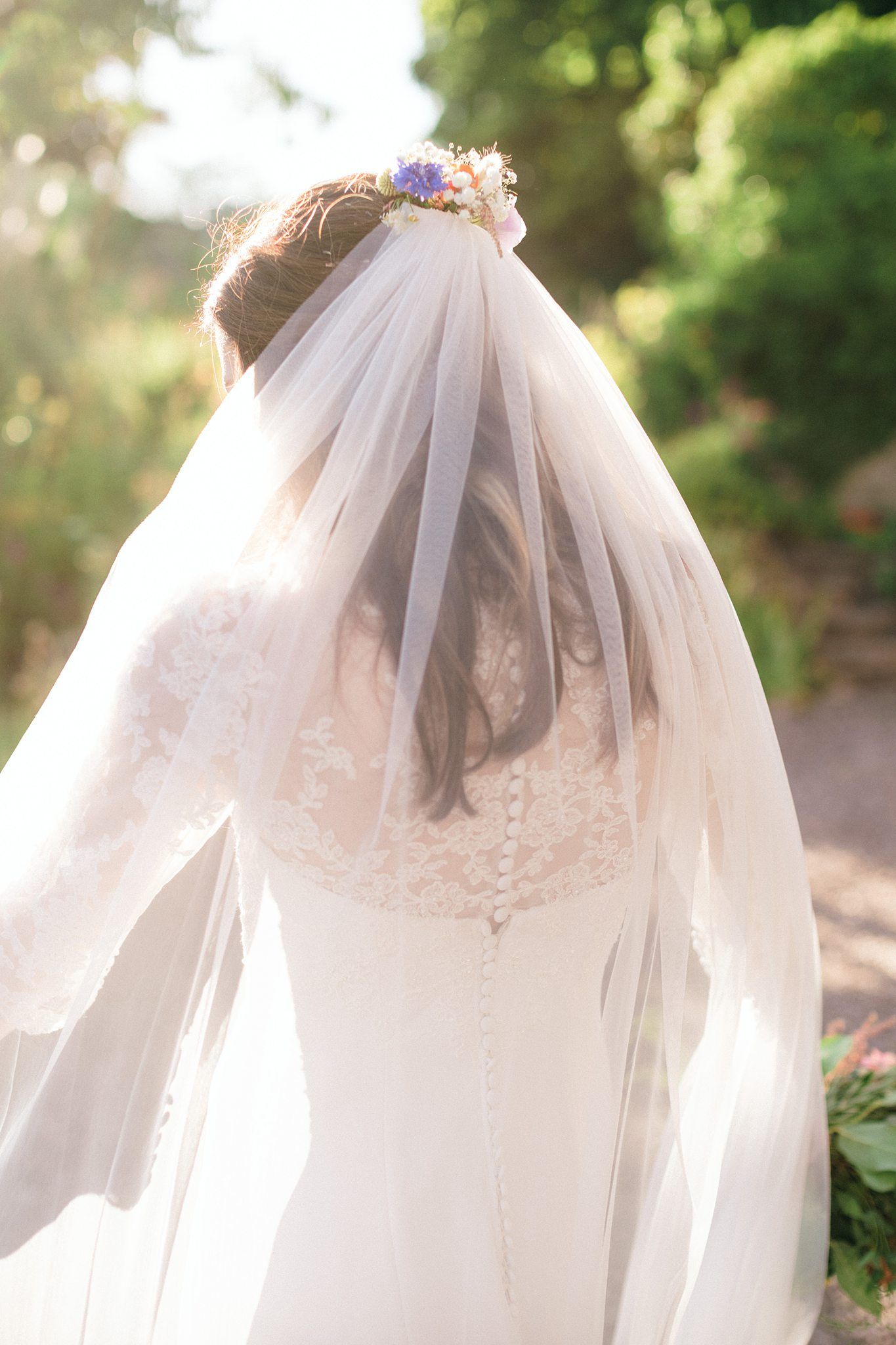Back view of bride wearing lace wedding dress and flowing white veil with small purple flowers in her hair at the Farm at South Cove.