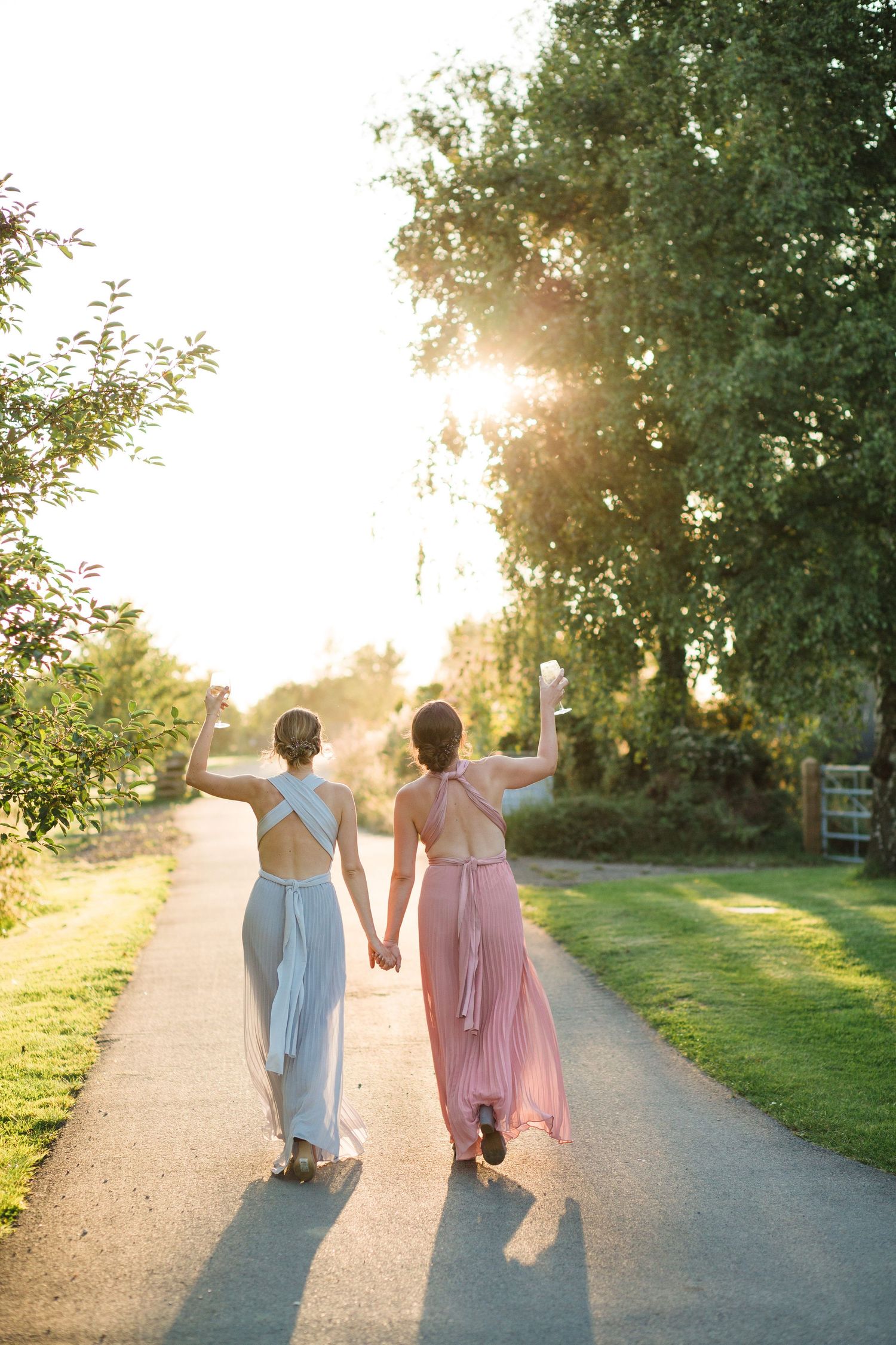 Two bridesmaids walk hand in hand on a sunlit path during golden hour with trees in the background at The Farm at South Cove, NS.