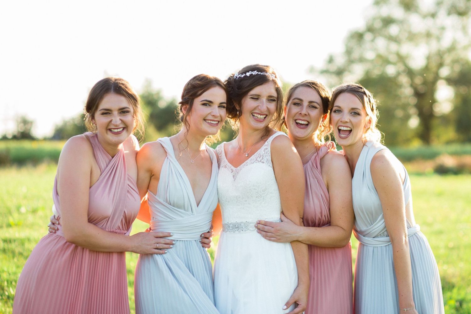 Colorful bridal party wearing pink and blue dresses stands together laughing in a sunny field during summer at the Farm at South Cove, NS.