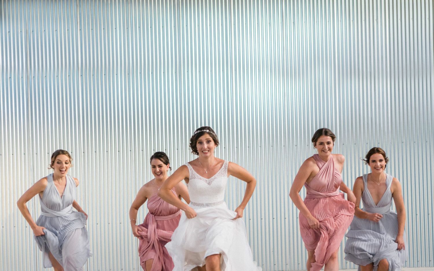 Bridal party runs playfully in front of a silver corrugated metal wall at Lunenberg's wedding venue the Barn at South Cove.