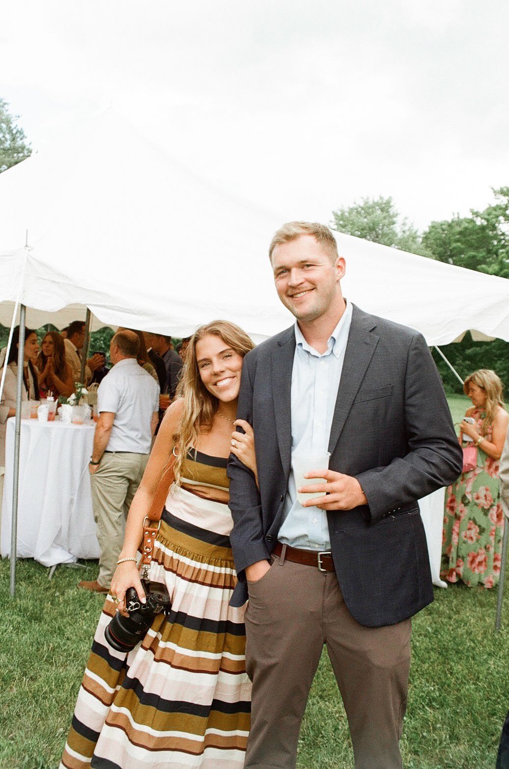 Smiling couple poses under white tent at outdoor garden party, woman in striped dress holding camera.