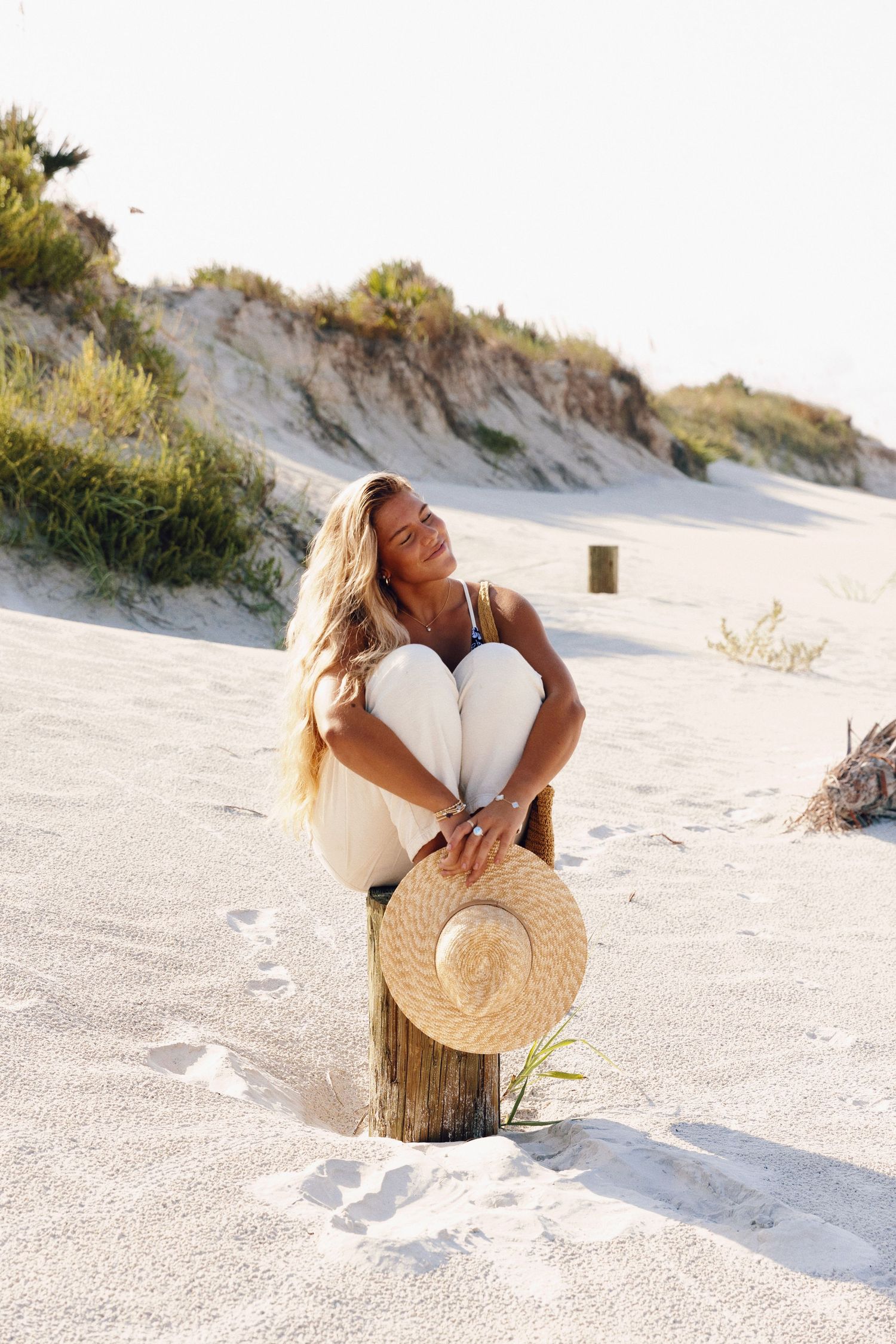 Person in casual attire holding straw hat while sitting on wooden post at beach.