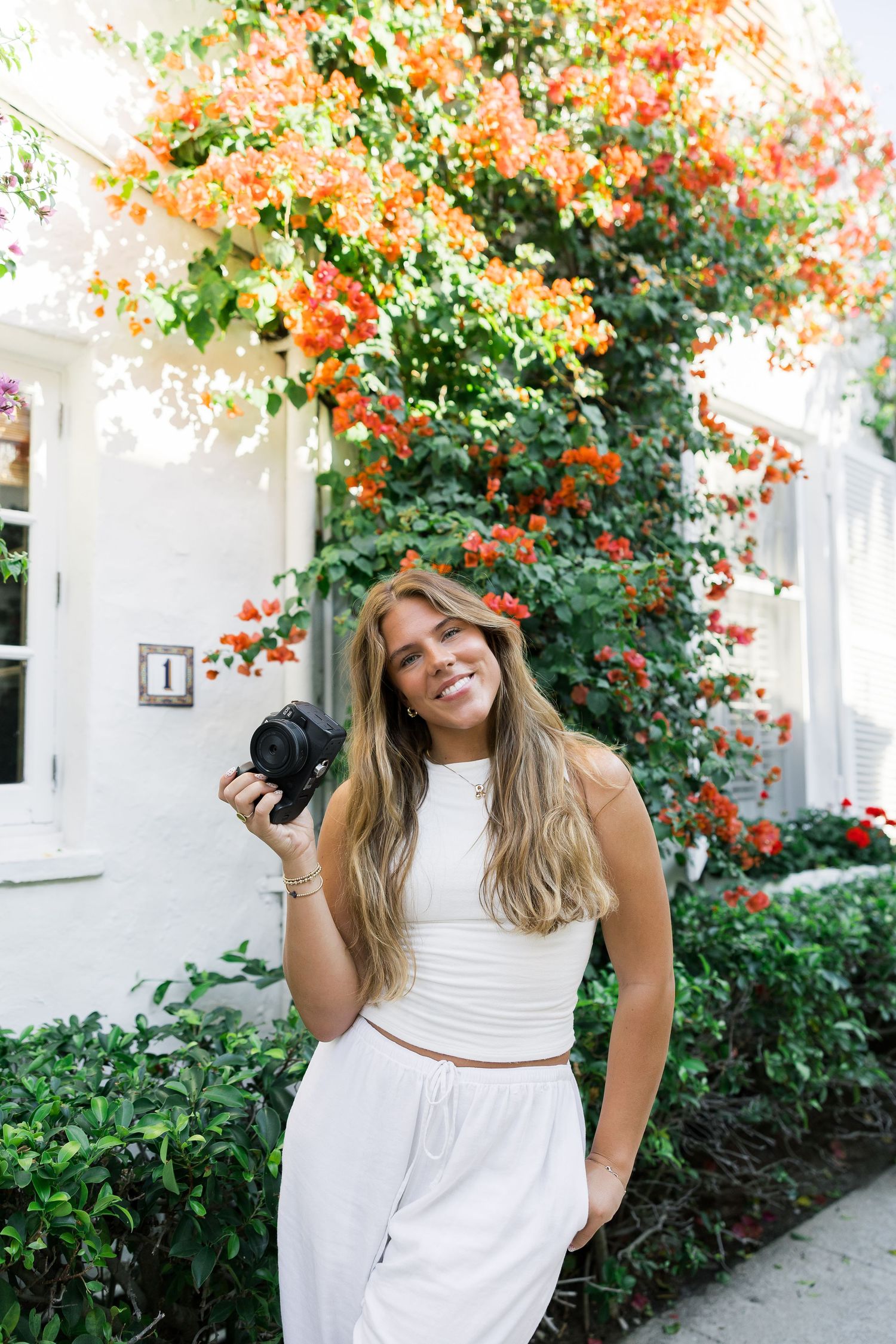 Smiling woman in white outfit holding a camera outdoors with vibrant orange bougainvillea flowers.
