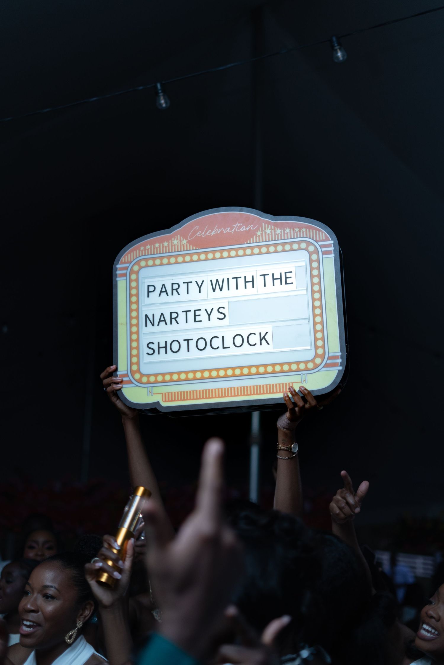 Silhouetted crowd holds up illuminated signs reading 'SHOTOCLOCK' and 'PARTY WITH THE NARTEYS' at a dark event venue.