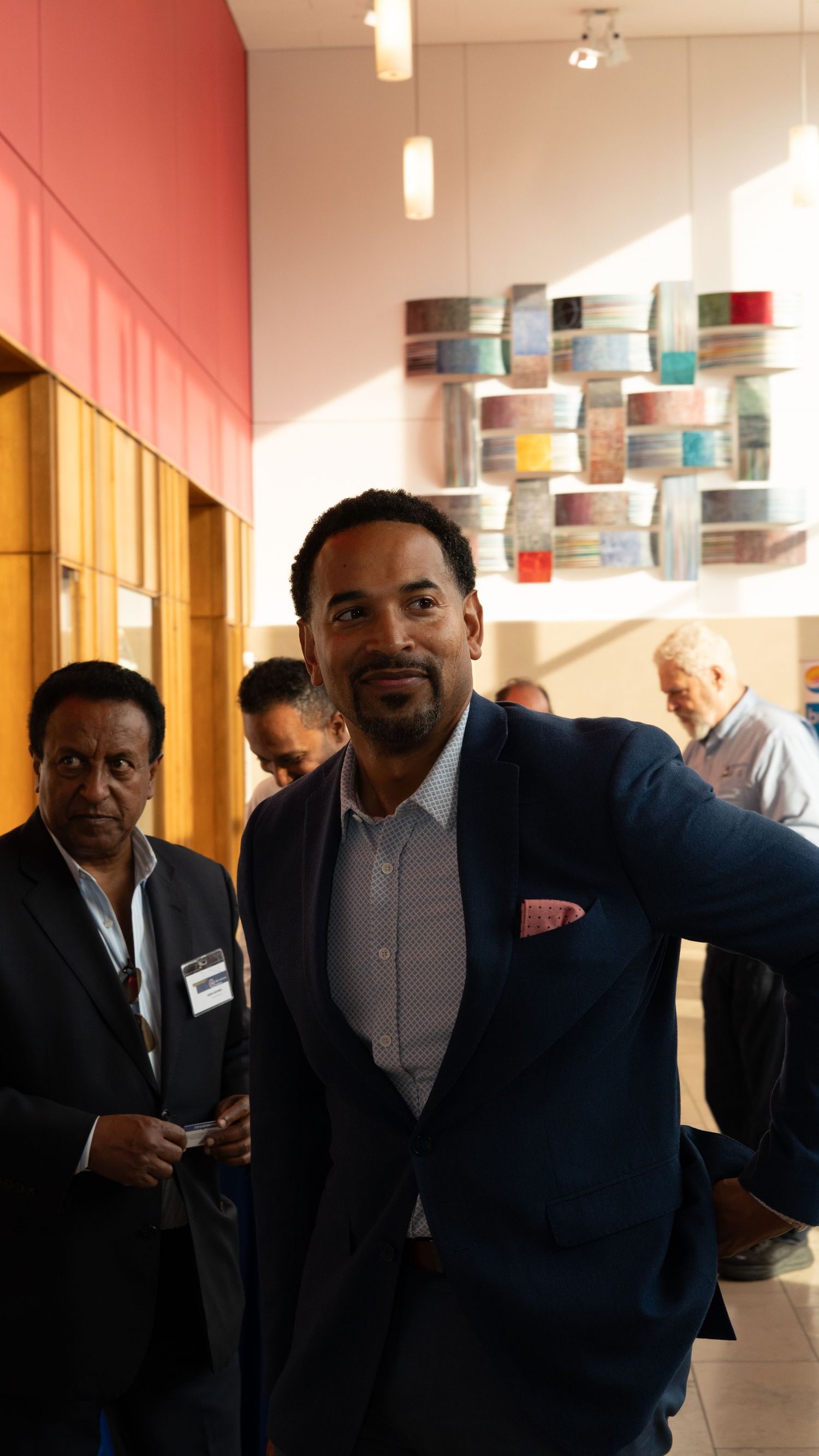 Professional man in navy suit with pink pocket square stands in modern lobby with colorful wall art.