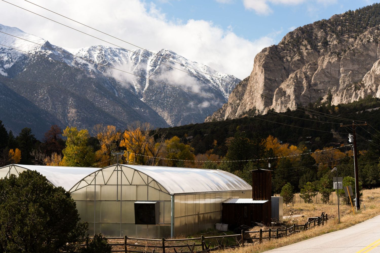 A greenhouse near a mountain road with snow-capped peaks and autumn foliage in the background.