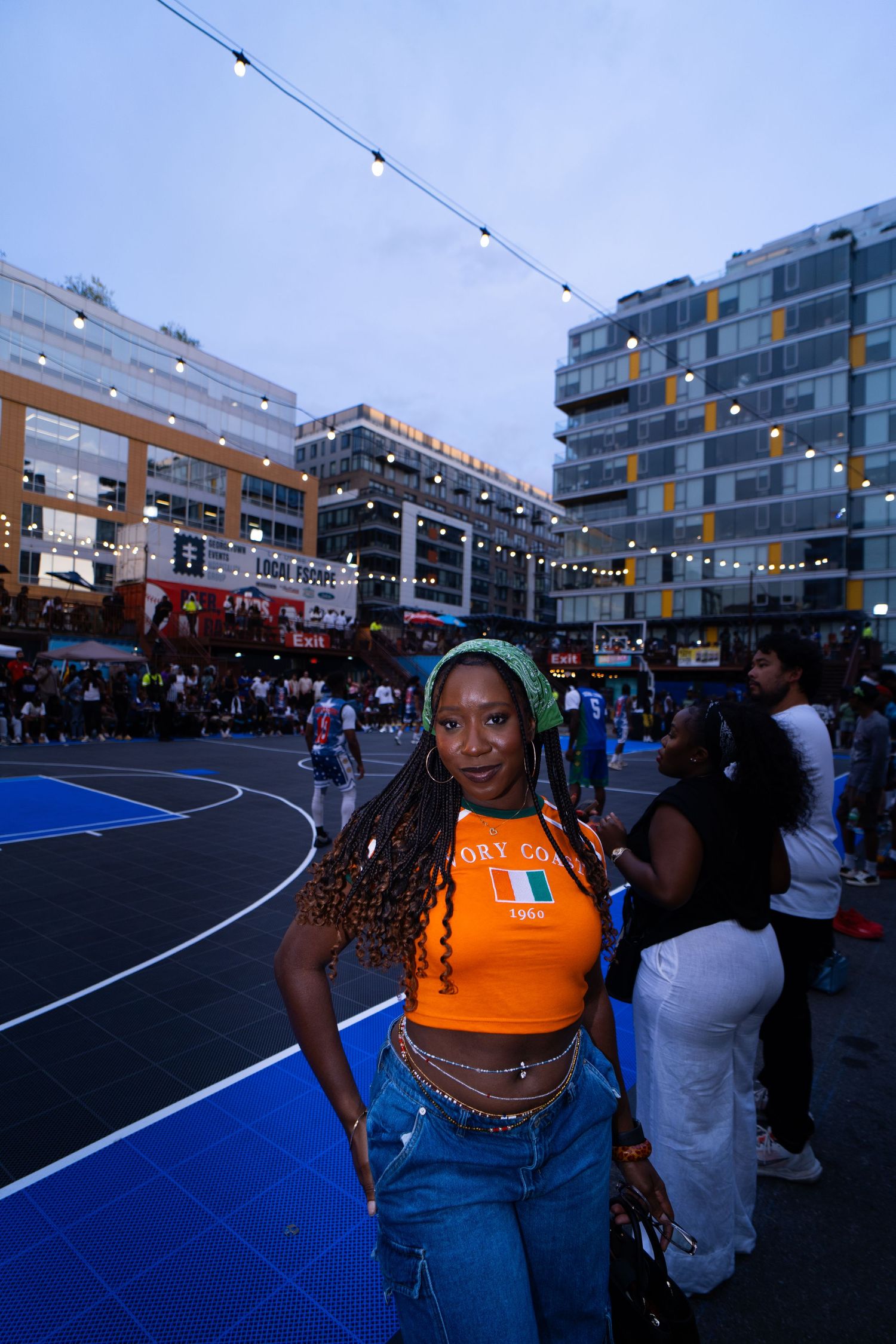 Woman in orange crop top and blue jeans poses on outdoor basketball court at dusk with string lights.