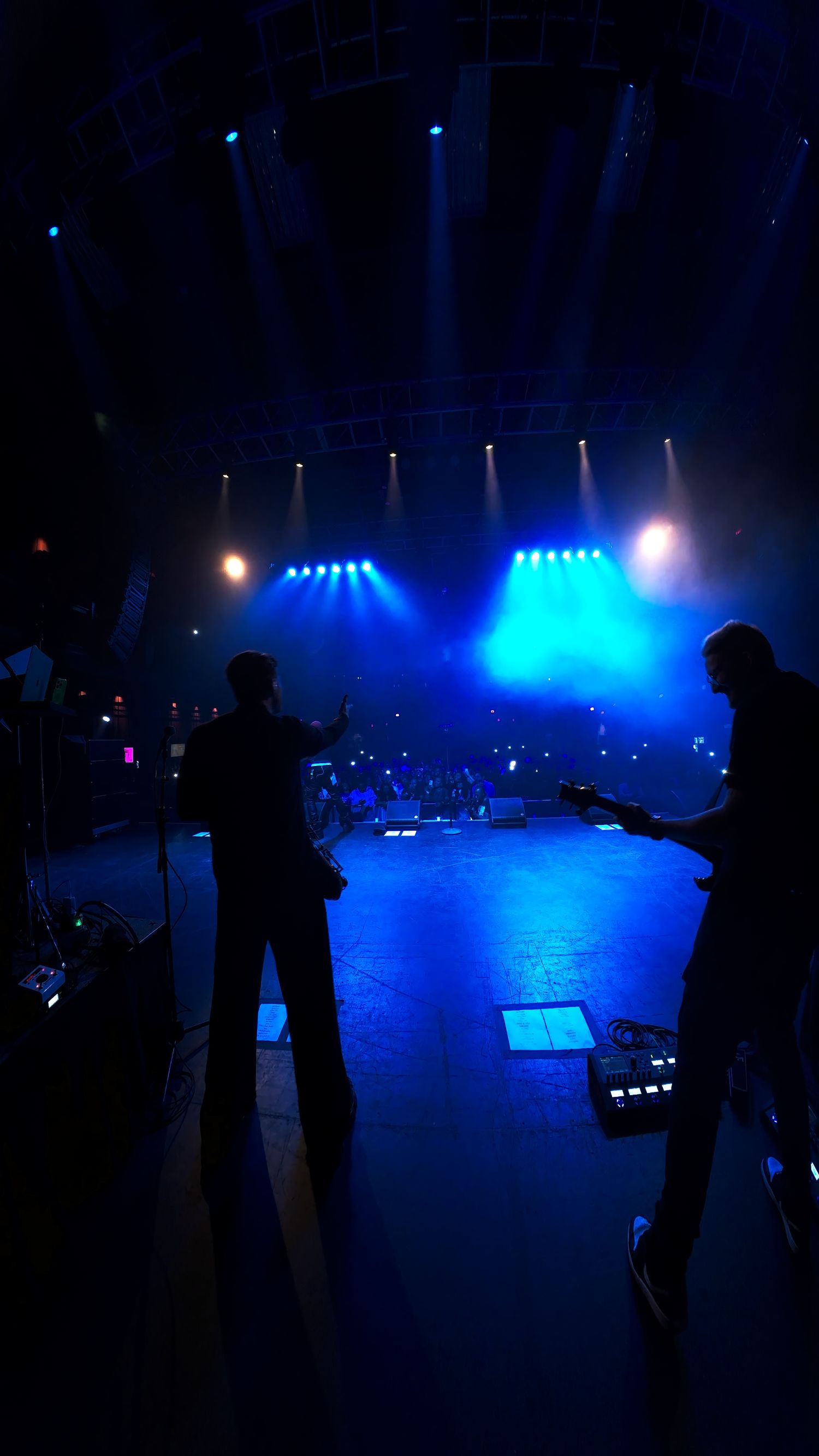 Concert stage setup with dramatic blue lighting and silhouetted figures against illuminated backdrop.