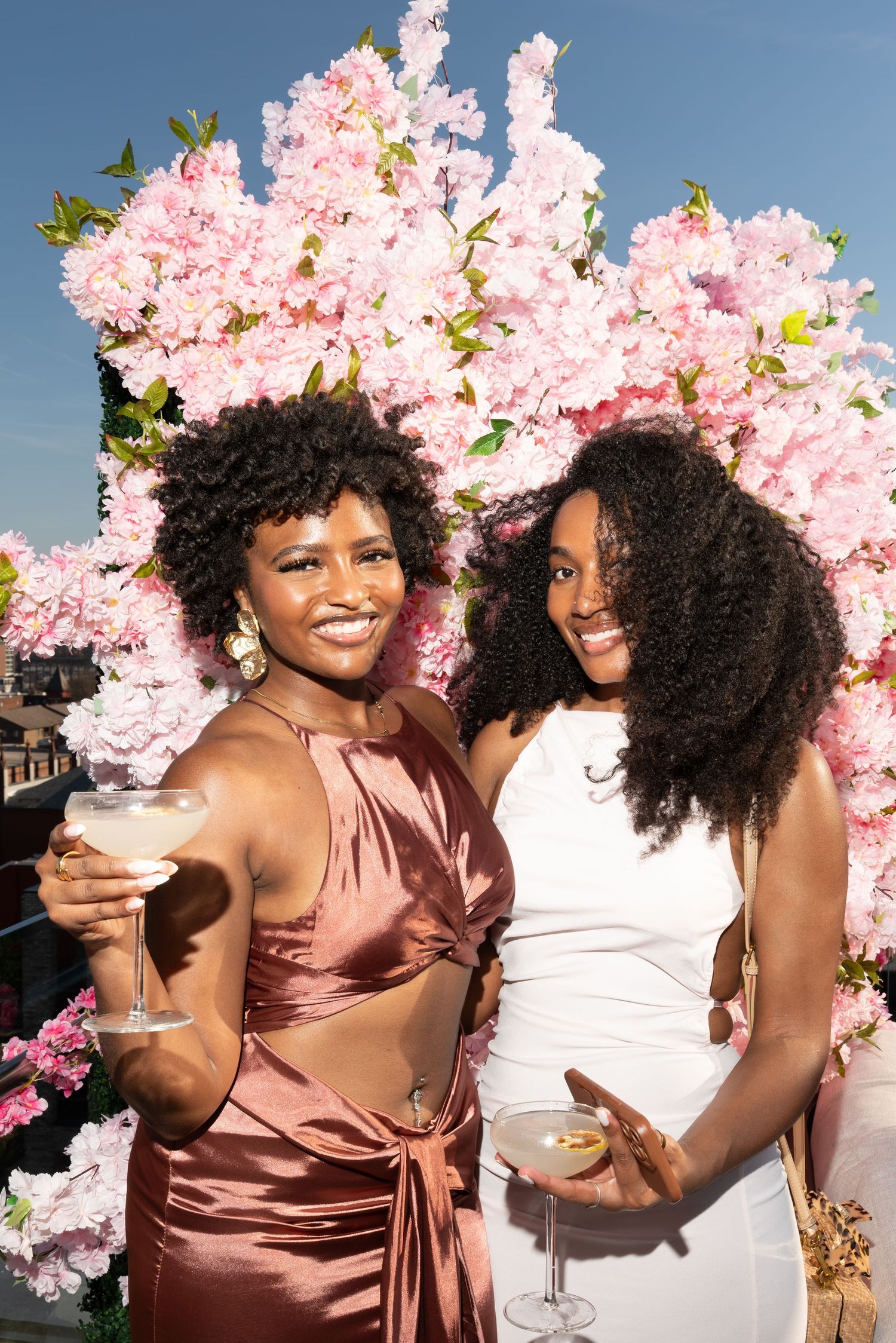 Two women smiling and holding cocktails in front of a pink floral backdrop at an outdoor event.
