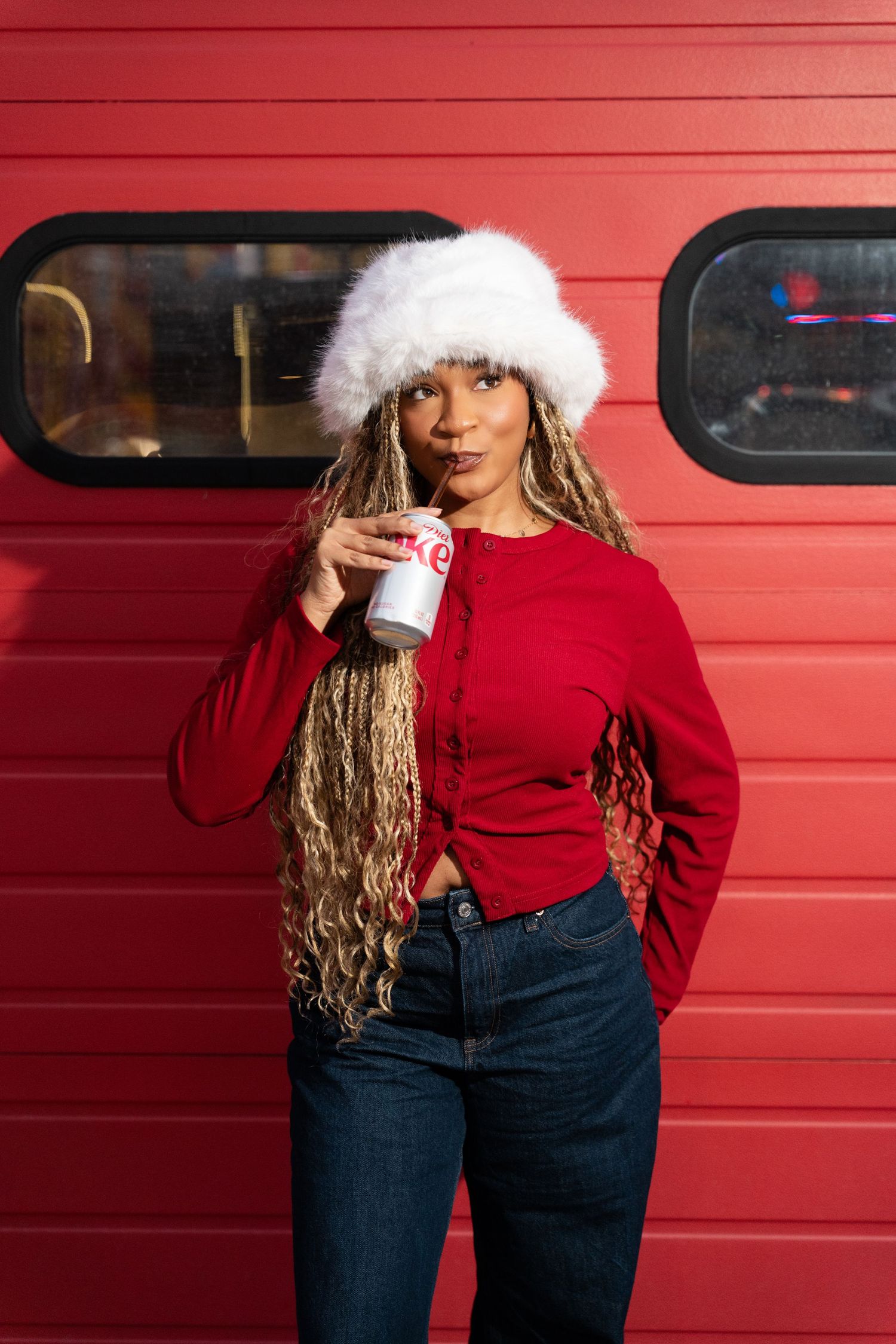 Person in red sweater and jeans poses against a red garage door wearing a white winter hat.