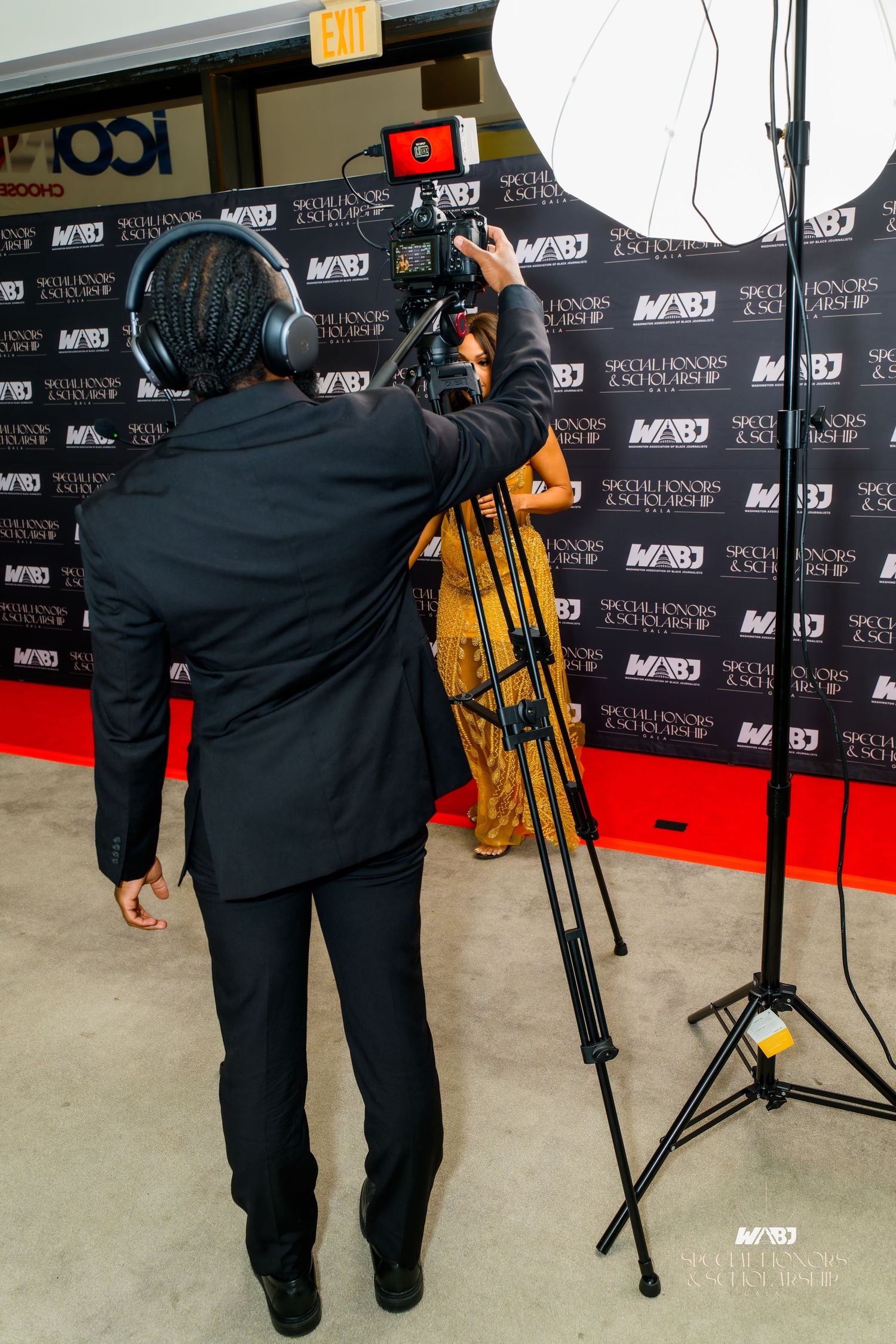 Camera tripod stands in front of black and red step-and-repeat backdrop at media event.