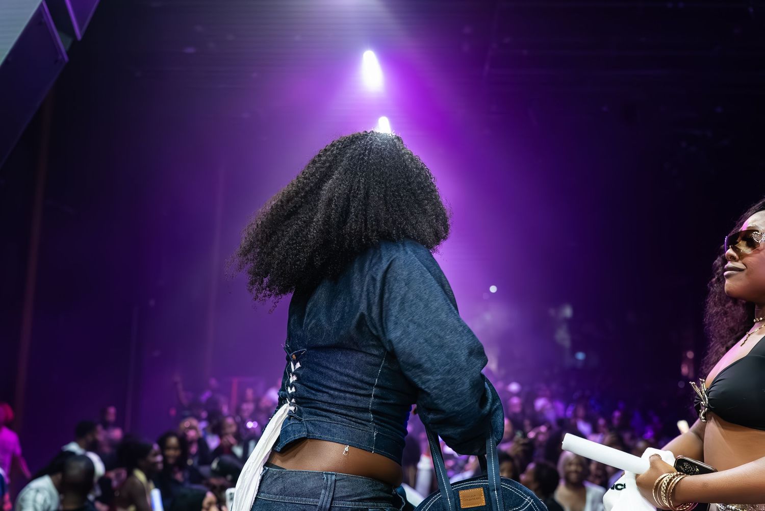 Woman with curly hair in denim outfit faces a purple-lit concert stage crowd at a live music event.