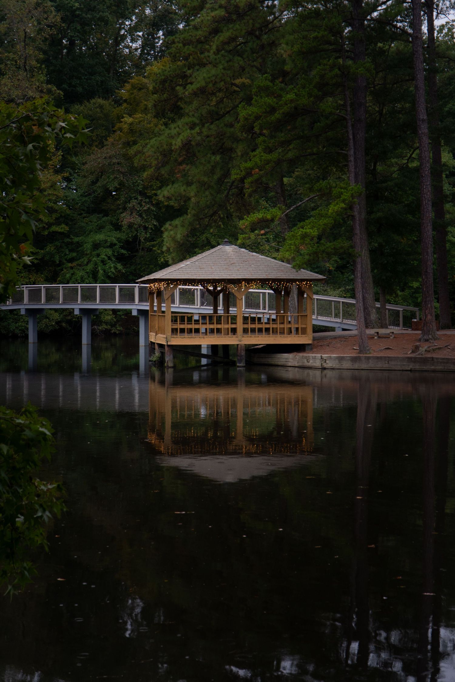 Wooden dock and gazebo structures reflected in calm lake waters surrounded by trees at dusk.